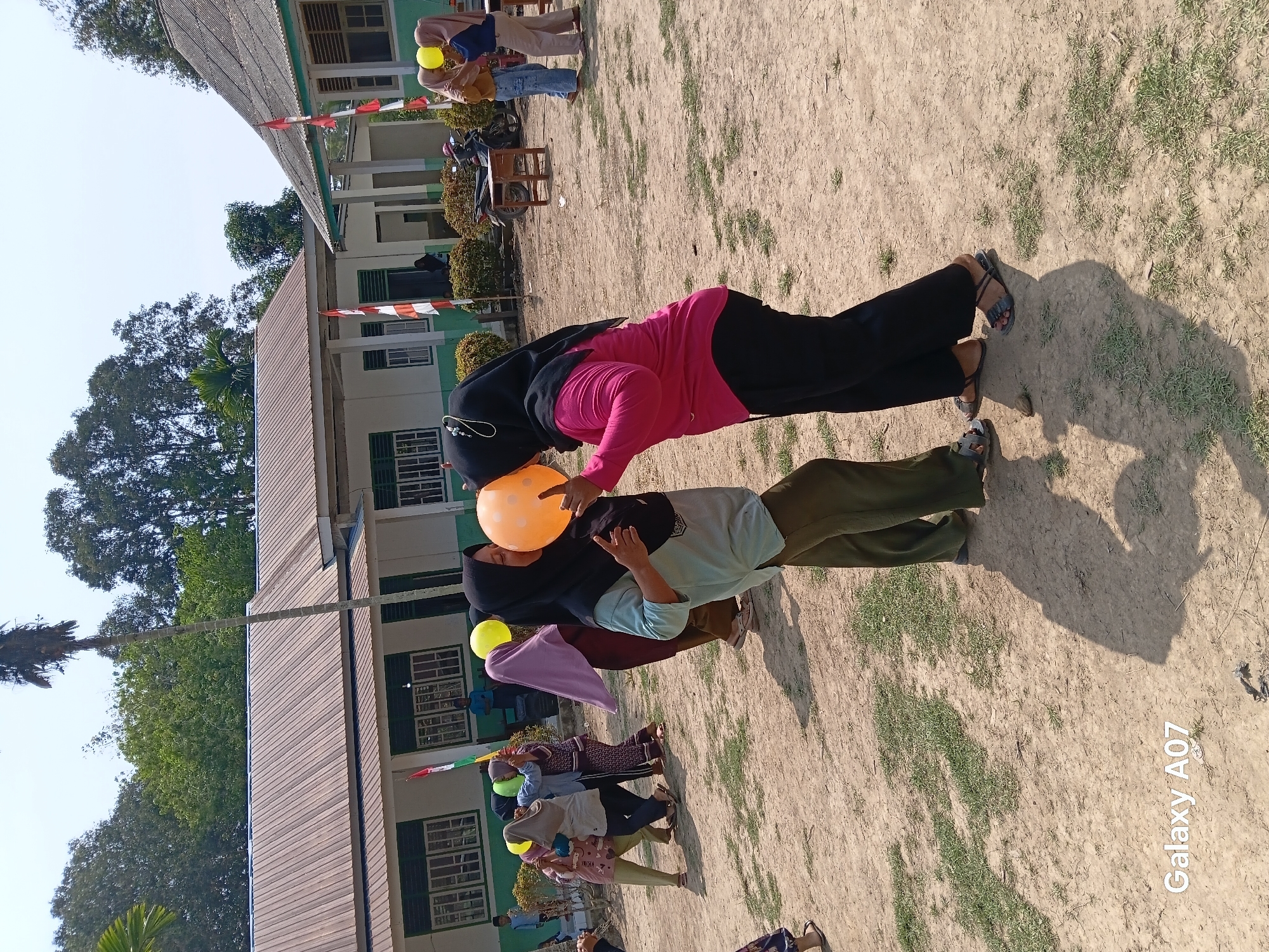 In Margo Rukun, Indonesia, two women wearing hijabs are engaged in a balloon-balancing game outdoors. One woman in a light blue shirt and dark green pants is holding an orange balloon between her face and the face of the other woman, who is wearing a pink shirt and black pants. The second woman appears to be biting the balloon. Several other women, also in hijabs, are visible in the background, some holding balloons. The scene takes place on a dirt ground with sparse grass, in front of a school building with white walls and a metal roof. Indonesian flags are displayed on poles near the building. The sky is clear and blue, and the sun casts shadows on the ground. The overall atmosphere suggests a lively outdoor event or competition.