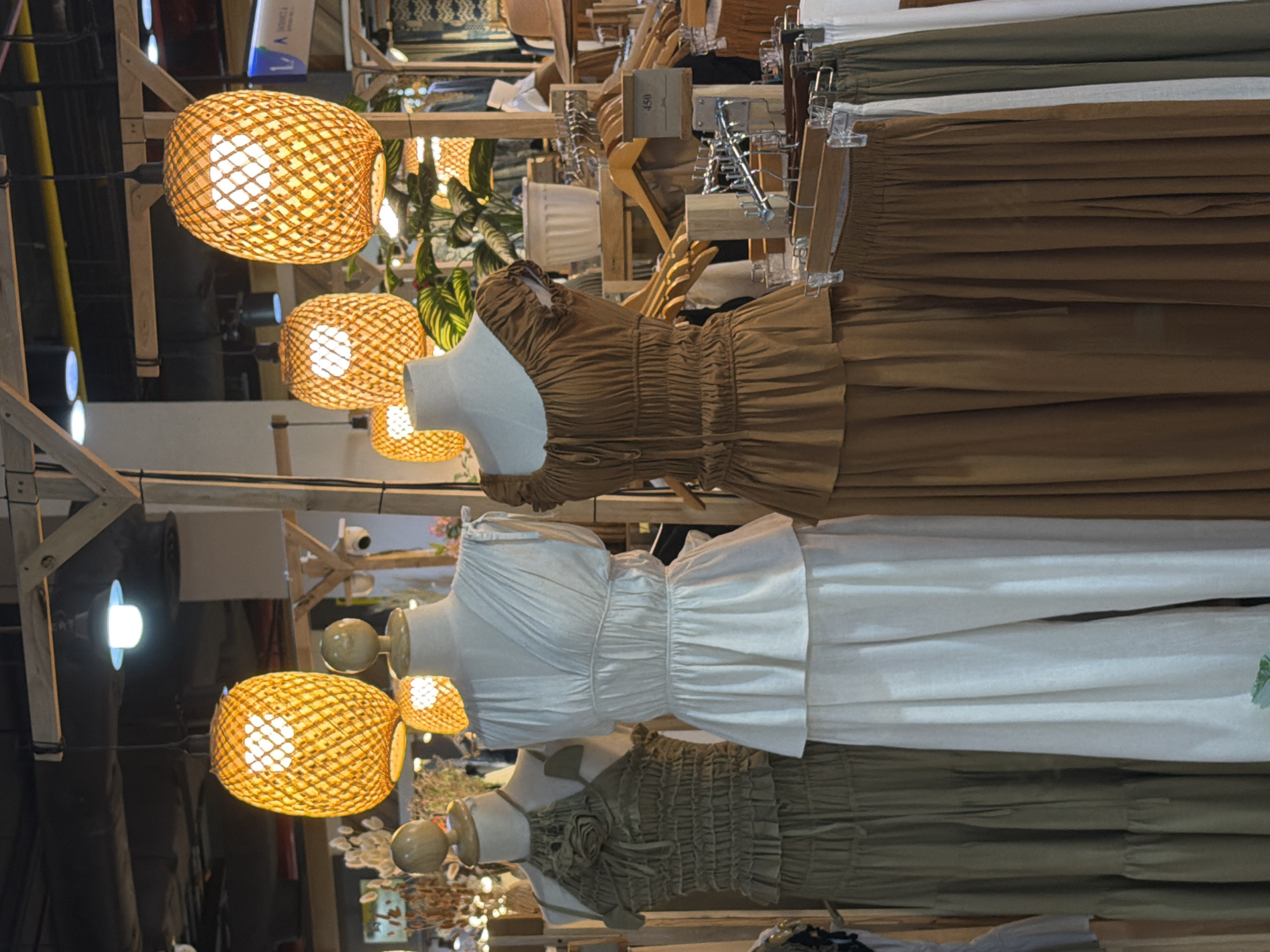This detailed image depicts a clothing display within an indoor market or retail space, likely located in Bangkok, Thailand. The scene is dominated by three female mannequins showcasing light, earthy-toned apparel, presented under warm, ambient lighting.
The leftmost mannequin wears a long, olive-green dress featuring a ruched, textured bodice with a halter neck and a prominent, layered fabric detail resembling a flower on the chest. The skirt of the dress is tiered. The central mannequin displays a cream-colored two-piece outfit consisting of a V-neck, short-sleeved top with a ruched waistline and a subtle peplum hem, paired with wide-leg trousers. The rightmost mannequin is dressed in a rust-brown two-piece ensemble, including a blouse with short, ruffled sleeves, a gathered neckline with a drawstring tie, and a ruched bodice with a peplum hem, complemented by a matching tiered skirt.
Above the mannequins, several distinctive oval-shaped hanging lamps with woven bamboo or rattan shades emit a warm, orange glow, contributing to a natural or bohemian aesthetic. These lamps are suspended from wooden beams that form part of the display structure. In the background, additional clothing items, primarily skirts and trousers in similar brown, olive, and white tones, are visible on wooden hangers on metal racks. A small brown price tag, clearly showing "450 THB," indicates the price in Thai Baht, confirming the location as Thailand. A potted plant with large, green leaves is positioned behind the central mannequin, and various other store elements, including blurred human figures, more overhead lighting, and general market infrastructure, can be seen in the distant background. The overall presentation suggests a focus on comfortable, natural fiber clothing within a charming, artisan-style retail environment.