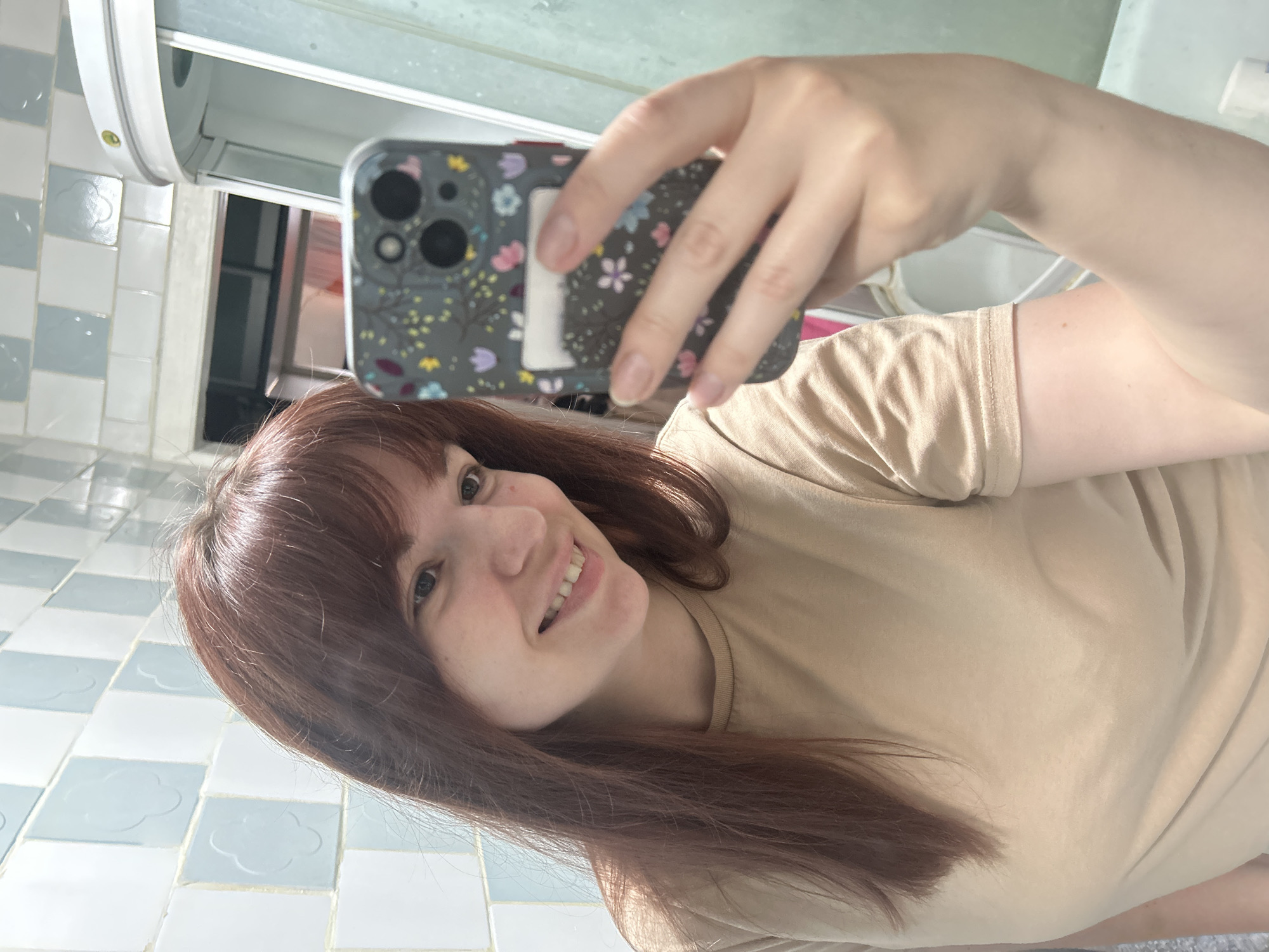 A woman with shoulder-length auburn hair is taking a selfie in a bathroom. She is holding a smartphone with a floral-patterned case, positioned slightly above her head and angled downwards. The woman is smiling and wearing a light brown t-shirt. The bathroom has light blue and white tiled walls with a floral pattern. The image does not provide enough information to determine the exact location in the city of Фоки, Russia.