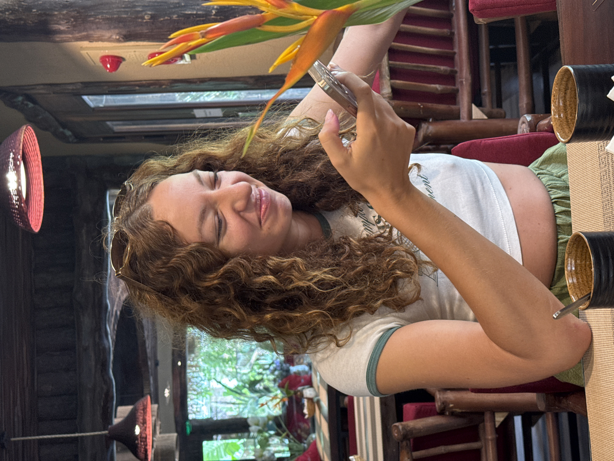 A young woman with curly brown hair is seated at a table in what appears to be an indoor restaurant. She is wearing a white crop top with a green trim and green pants. She is holding a mobile phone in her right hand and looking down at it, smiling. Her left arm is resting on the table. In the foreground, there are two dark brown cups with a textured pattern. Behind her, a bright orange and yellow flower is visible. The background shows wooden chairs and tables, as well as a window with greenery visible outside, suggesting a tropical or lush setting. The lighting appears to be warm and ambient.