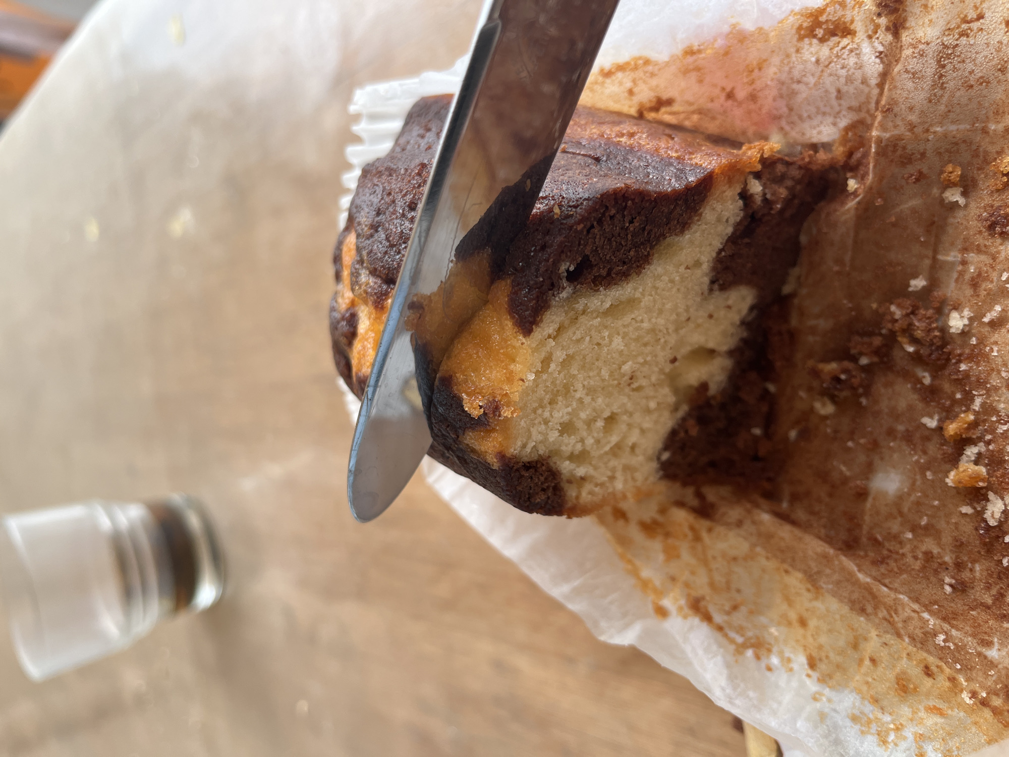 A slice of marbled cake is being cut by a knife on a wooden table. The cake is on a white paper wrapper, with crumbs scattered around. In the background, a glass of dark liquid, possibly coffee or espresso, is visible. The scene is likely set at a cafe or restaurant in Palavas-les-Flots, France.