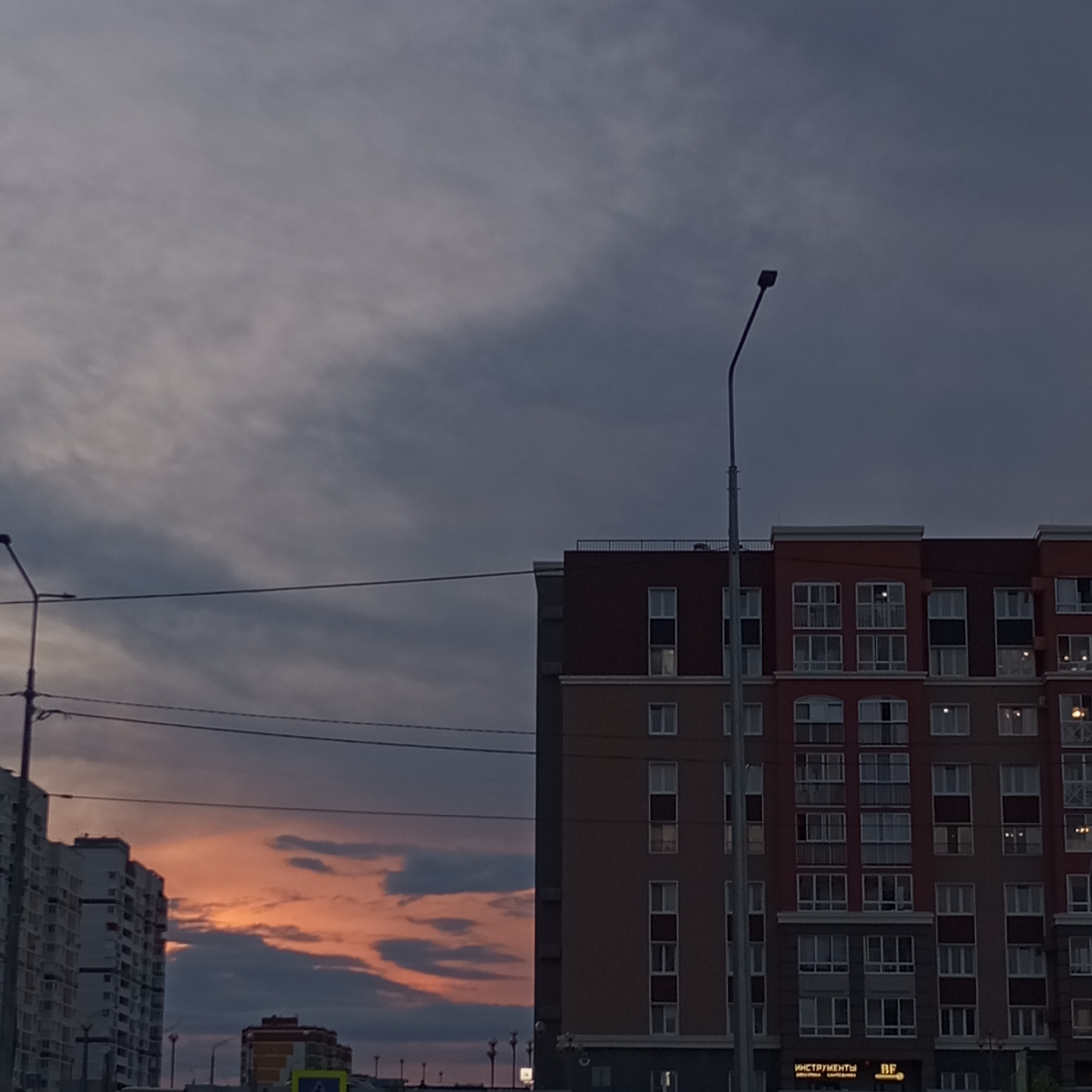 This is a low-angle, full-shot photograph of a cityscape at dusk, featuring a modern apartment building on the right and a taller residential building on the left. The sky is filled with dramatic, cloudy patterns, illuminated by the warm hues of a sunset or sunrise, with shades of orange and pink visible through the darker, overcast clouds.
The apartment building on the right is a multi-story structure with a distinctive facade, featuring alternating bands of darker red and beige. Numerous windows, many with balconies, are visible across its surface. Some windows are illuminated from within, suggesting occupancy. A street lamp is prominently positioned in the foreground, to the left of this building, with its arm extending upwards and to the right.
The building on the left is a much taller, white-paneled residential block, appearing more utilitarian in design. Its vertical structure dominates the left side of the frame.
In the background, other smaller buildings and urban infrastructure are visible, including streetlights and what appear to be overhead wires. A sign with Cyrillic lettering is visible on the lower part of the right-hand apartment building. The overall impression is of a contemporary urban environment during a time of changing light.
The image does not contain any discernible people or overt actions. The focus is on the architecture and the atmospheric sky. The scene is set in the city of Pervomaysky, Russia.