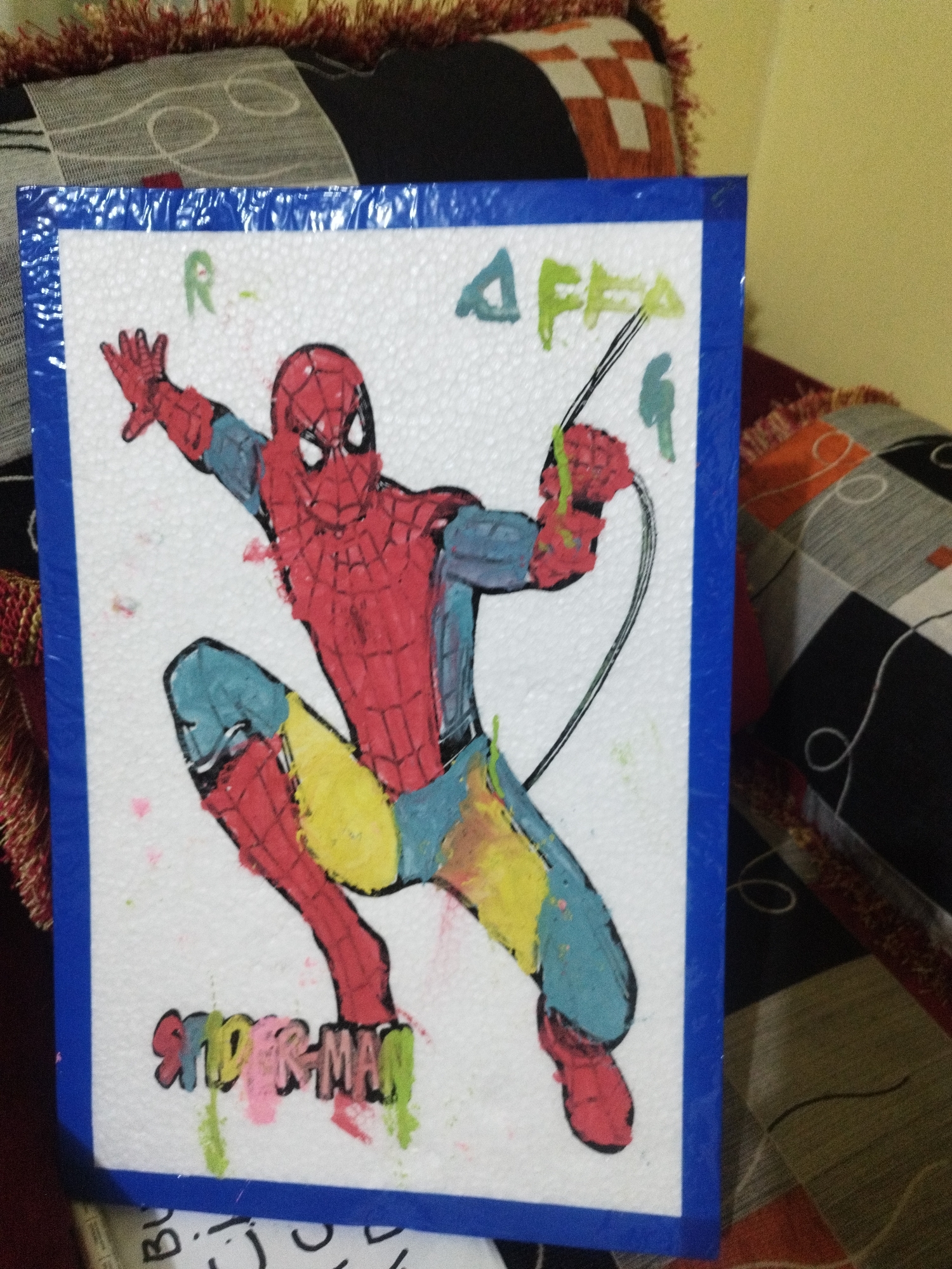 This is a close-up shot of a framed drawing of Spider-Man, likely a child's artwork, placed on a patterned surface, possibly a cushion or rug. The drawing is on a white, textured material, possibly styrofoam, and is bordered by a blue frame. Spider-Man is depicted in a dynamic pose, with his webs extending upwards. The colors used are vibrant, including red, blue, yellow, and green. Text is visible above and below the drawing, with "R" and "AFFA" written in what appears to be green marker, and "SPIDER-MAN" written in various colors below. The background is blurred, but suggests a domestic setting with patterned fabrics and a yellow wall. There is no visible information linking this image to Sumenep, Indonesia.