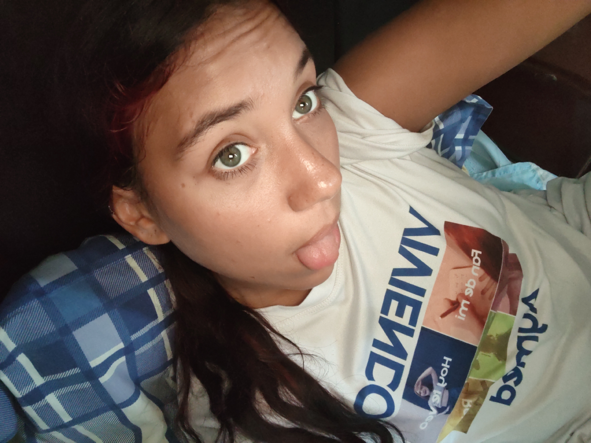 The image shows a close-up, high-angle selfie of a young woman with brown hair and green eyes. She is lying down, wearing a white t-shirt with blue and red graphics and a blue and white plaid blanket or shirt underneath. Her tongue is sticking out. The background is dark, suggesting an indoor setting. The photograph was taken in Piloncito, Venezuela.