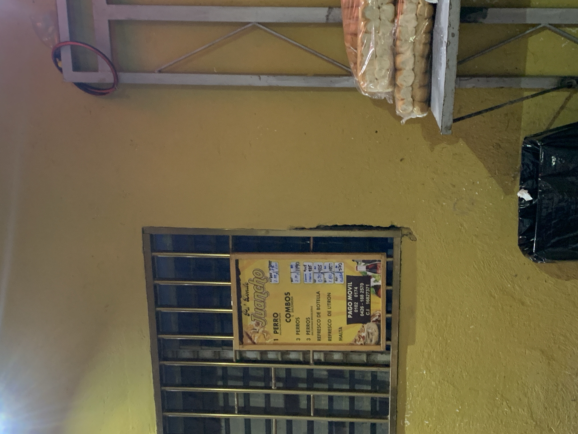 The image captures a static outdoor scene at what appears to be a small food stall or establishment at night, indicated by the artificial lighting and overall dimness. The primary background is a textured, light yellow wall.
At the top of the frame, there's a grey metal structure, possibly part of an awning or shelf support. From this structure, several clear plastic bags filled with small, light-colored bread rolls are hanging, suggesting they are for sale. To the right, a dark, possibly black, object resembling a trash bag or container is partially visible. On the bottom left, a bright light source creates a significant flare, obscuring part of the wall.
A prominent feature is a menu or sign, framed in wood, hanging within a barred window. The text on the sign is in Spanish. It names the establishment "De Esquina Juancho" and lists "COMBOS." Offerings include:
- "1 PERRO" (1 Hot Dog) with "SALCHICHA COMPLETA" (complete sausage), priced at 2.10 and 1.90.
- "3 PERROS" (3 Hot Dogs) with "SALCHICHA," priced at 905.
- "3 PERROS" (3 Hot Dogs) with "SALCHICHAS BRASILERAS" (Brazilian sausages), priced at 485.
Drinks available are "REFRESCO DE BOTELLA" (Bottled soda) at 0.75 and 425, "REFRESCO DE LITRON" (Liter soda) at 1.80 and 10, and "MALTA" (Malt drink) at 0.65 and 370. The sign also provides "PAGO MOVIL" (Mobile Payment) details, including phone numbers (0102 0174, 0426-188 2570) and a C.I. (ID number) 16827371. The prices, especially the larger ones, are likely in an older currency or smaller units, while the smaller numbers might represent current prices in USD or a similar strong currency often used in Venezuela for daily transactions. The overall impression is a modest, local eatery offering hot dogs and drinks, catering to customers with mobile payment options.