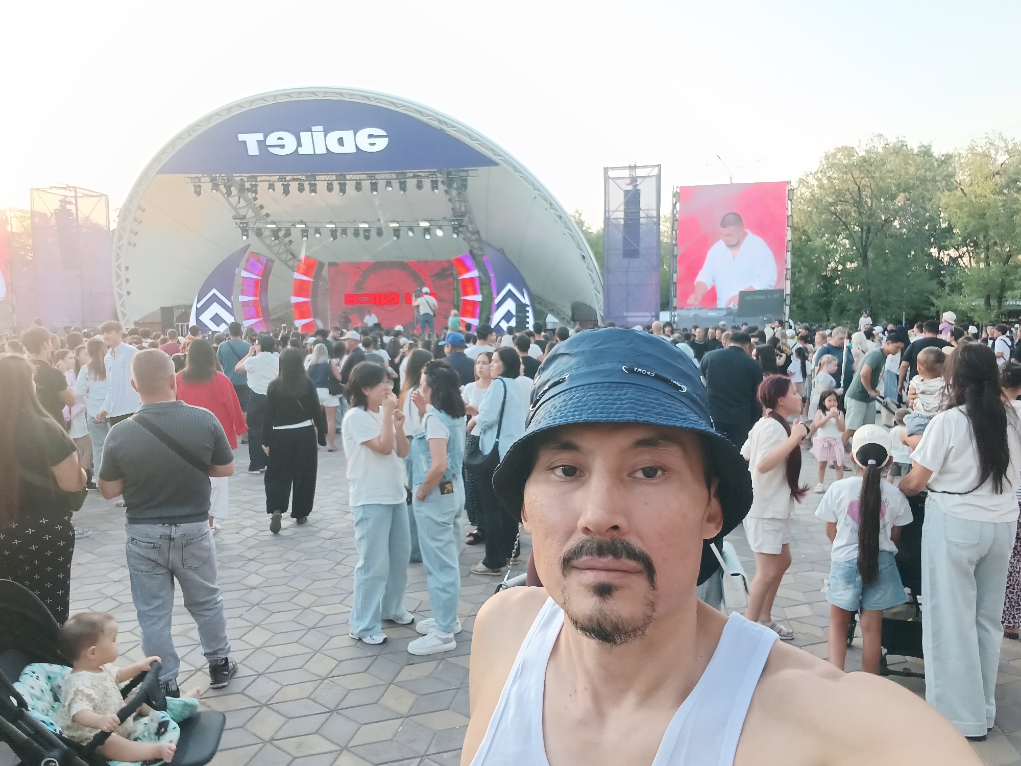 The image captures a selfie taken by a man with a mustache and goatee, wearing a white tank top and a blue bucket hat. He is standing in the foreground, facing the camera. Behind him, a large crowd of people is gathered, suggesting an outdoor event or festival. A stage with a large curved canopy is visible in the background, featuring bright lighting and a screen displaying red graphics. To the right of the stage, another large screen shows a man in a white shirt. The ground is paved with interlocking geometric tiles. In the lower left corner, a baby in a stroller is visible. The scene appears to be set in a public space with trees and an open sky overhead. The sign above the stage, partially obscured, reads "TƏLİGE" in reverse. The overall atmosphere suggests a lively event taking place in an urban park or public square, possibly in Qarağandı city administration, Kazakhstan, given the potential presence of a large public gathering area and stage setup typical for such locations.