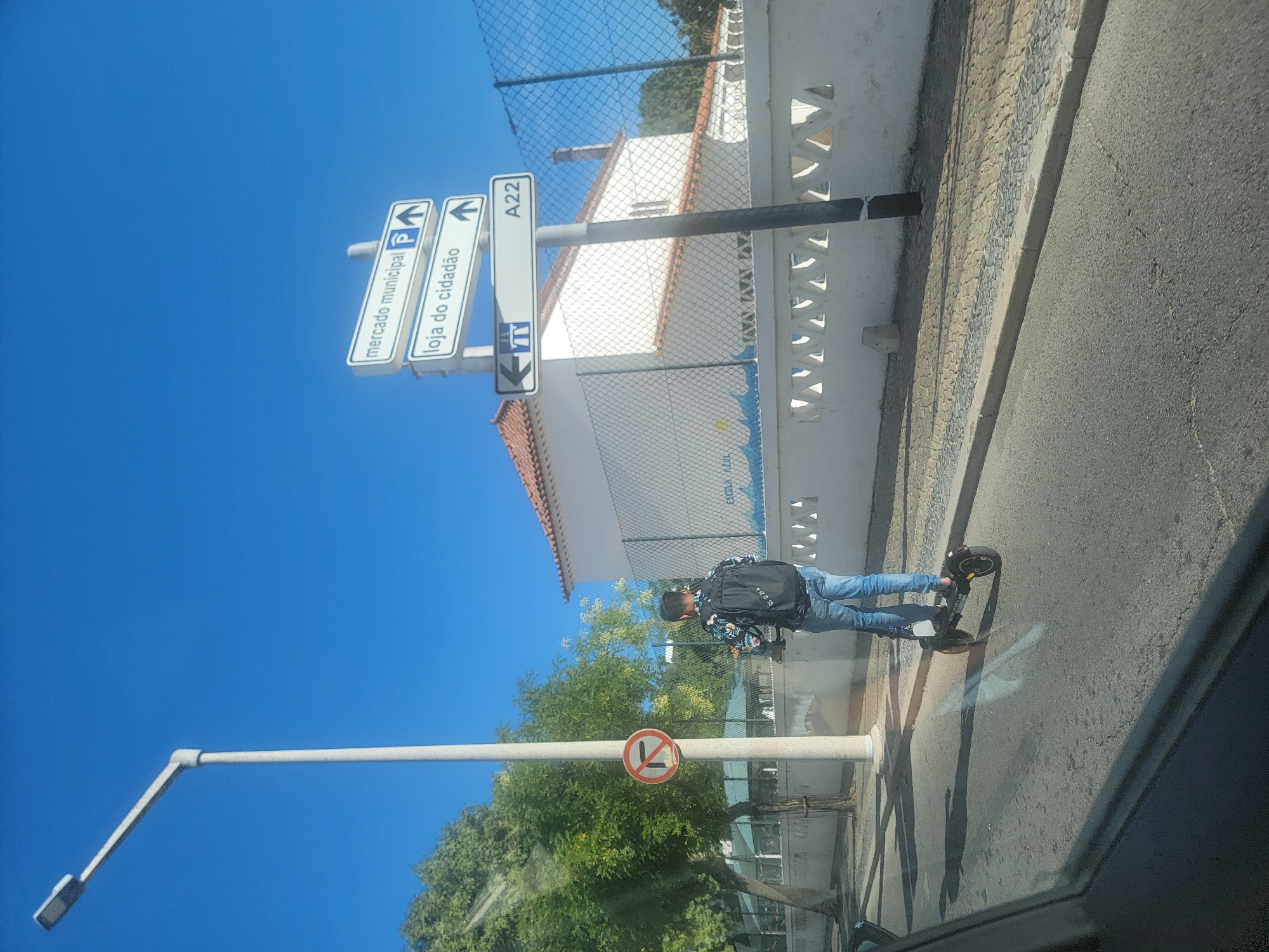 A young person is riding an electric scooter on a street in Faro, Portugal. The person is wearing jeans and a patterned shirt, and carrying a backpack. The street is paved with asphalt, and there is a sidewalk with cobblestones. In the background, there is a white building with a red tiled roof, a white decorative wall, and a chain-link fence. There are trees and a lamppost on the left side of the image. A traffic sign indicating "mercado municipal" and "loja do cidadão" with arrows pointing forward, and another sign indicating A22 with an arrow pointing left are visible. A "no left turn" sign is also visible on a pole. The sky is clear and blue.