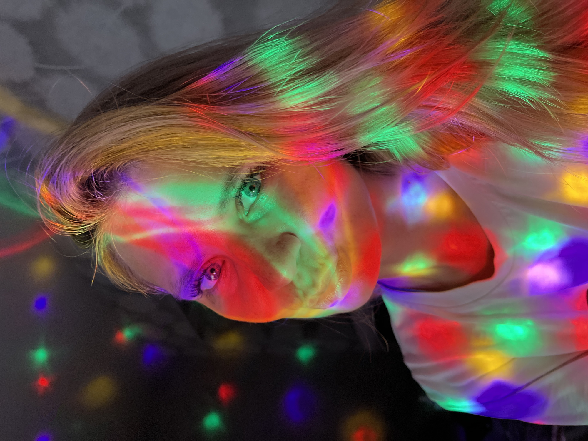 The image is a close-up portrait of a young woman, likely in her late teens or early twenties, illuminated by colorful disco lights. She is looking slightly to her left, with her eyes open and a subtle smile on her lips. Her hair, a shade of blonde, is caught in the light, showing streaks of pink, green, yellow, and red. The lights cast vibrant and abstract patterns across her face and her white t-shirt. The background is dark and out of focus, with more colored lights appearing as bokeh. There are no discernible landmarks or context to suggest a specific location within the city of Chaikovsky, Russia. The scene appears to be indoors, possibly at a party or gathering where a disco ball or similar light effect is present.
