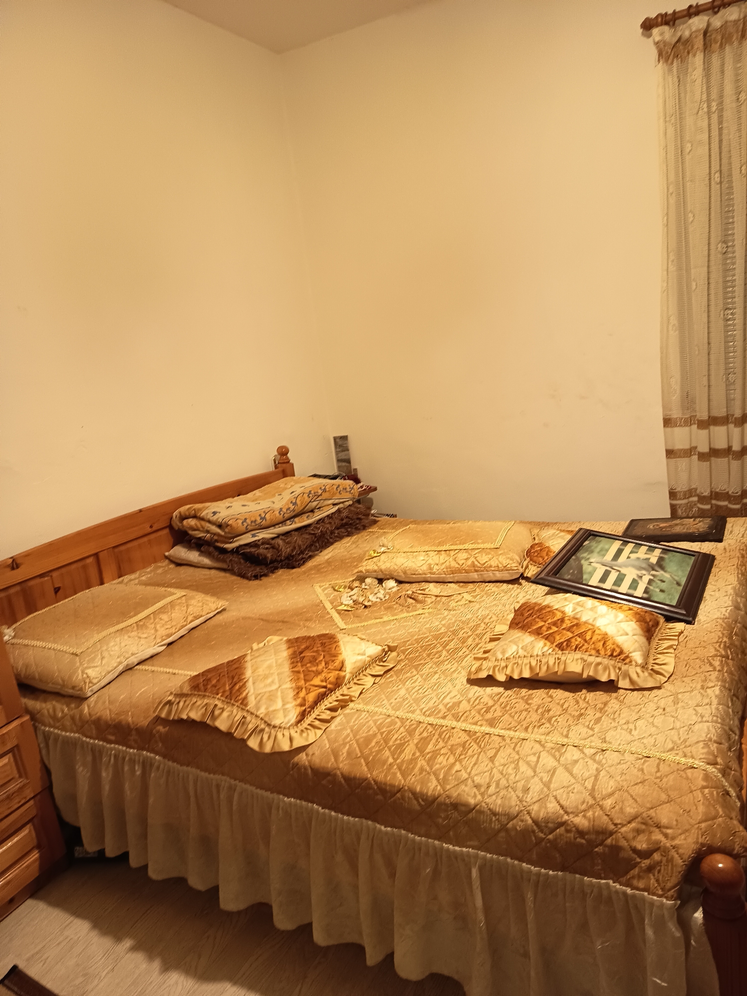 A bedroom features a double bed with a golden-quilted bedspread and matching pillows. Two framed pictures and some decorative ornaments are arranged on the bed, along with some folded blankets stacked at the head of the bed. A wooden headboard is visible, and light-colored curtains hang to the right of the bed. The walls are painted in a neutral color. The location is Unknown, Unknown.