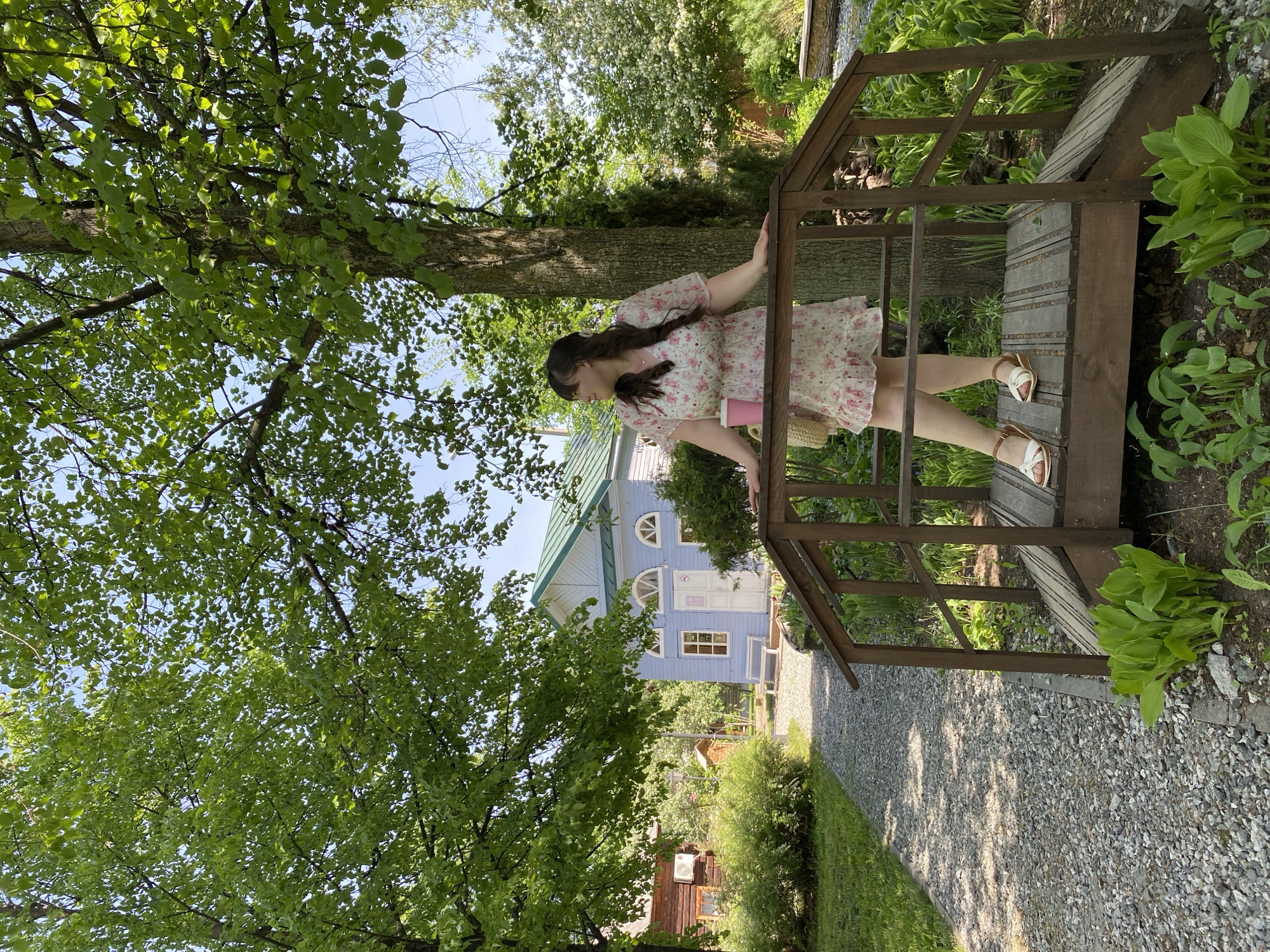 A young woman in a floral dress and sandals is standing on a small wooden bridge in a garden. She is holding a pink cup in her right hand and leans against the bridge's railing. The bridge spans a gravel path. Behind the woman is a blue house with a green roof and white trim. Lush greenery, including trees and shrubs, surrounds the scene. The location is likely in Votkinsk, Russia, given the prompt's specifications.