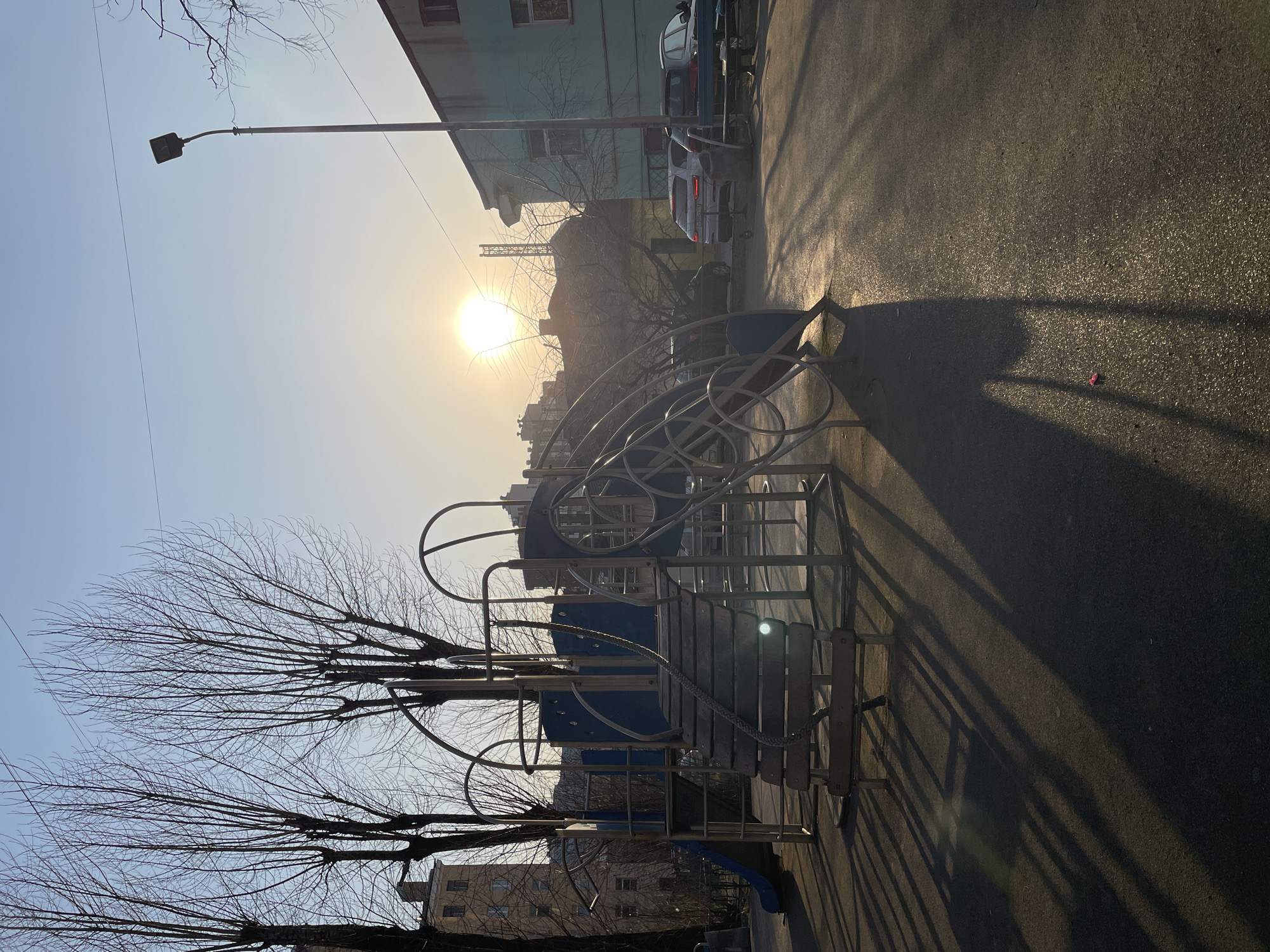 The image shows a playground structure, likely located in a residential area, with the sun prominently positioned in the background. The playground equipment appears to be made of metal and blue plastic. A street lamp stands tall to the left of the sun. Bare tree branches are visible framing parts of the scene, suggesting it may be late fall or winter. Several buildings are visible in the background, along with parked cars. The ground is paved, and the shadows of the playground structure cast long lines across the pavement. Given the architectural style of the buildings, the location is plausibly in Saint Petersburg, Russia.