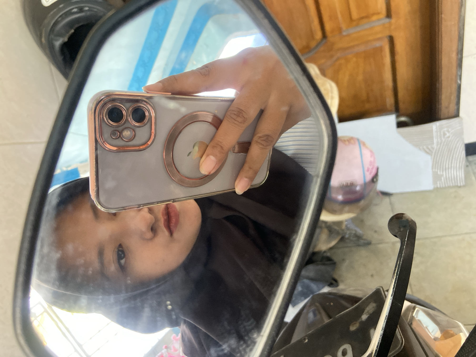A young woman wearing a black hijab is reflected in a motorcycle's side mirror. She is holding a smartphone with a rose gold case and a visible Apple logo. The reflection also shows part of a black motorcycle helmet in the upper right corner, and the woman's hand holding the phone. The background of the reflection appears to be a mix of indoor and outdoor elements, with wooden furniture and what looks like a window with horizontal blue and white stripes. The scene is set in the city of Gelam, Indonesia.