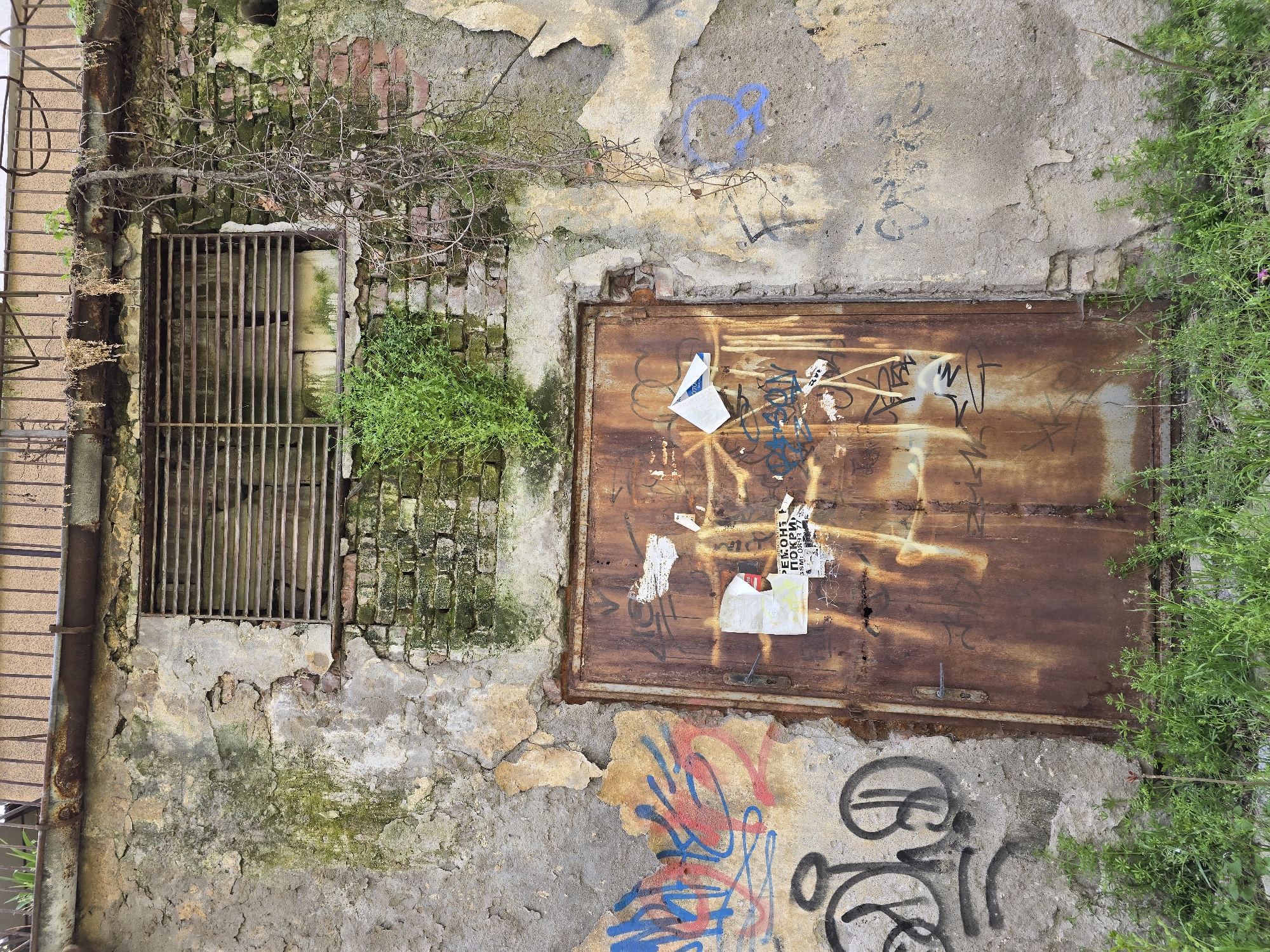 The image shows an exterior wall in an apparent state of disrepair. There's a barred opening or window to the left, covered with a rusty metal grate, and a metal door to the right, similarly rusted and marked with graffiti and affixed stickers. The wall itself is a mixture of brick and decaying plaster, with moss and vegetation growing on it. There is some graffiti on the lower part of the wall and also the metal door.