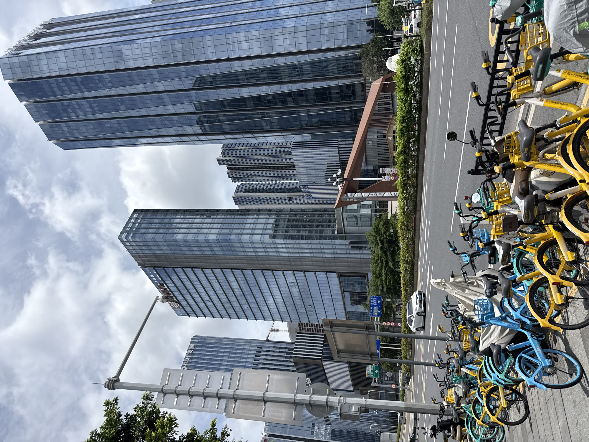 In Tianhe District, China, a row of colorful shared bicycles, predominantly blue and yellow, are parked along a sidewalk. Behind the bicycles, a multi-lane asphalt road runs horizontally, with a white sedan driving in the right lane. Further in the background, a dense collection of modern skyscrapers with glass facades dominates the urban landscape under a cloudy sky. A street lamp with a surveillance camera is positioned to the left of the frame, and a distinctive angular, reddish-brown structure with lights is visible in the mid-ground. Lush green trees and shrubs line the area between the sidewalk and the road, and a white, abstract sculpture is partially visible among the foliage.
