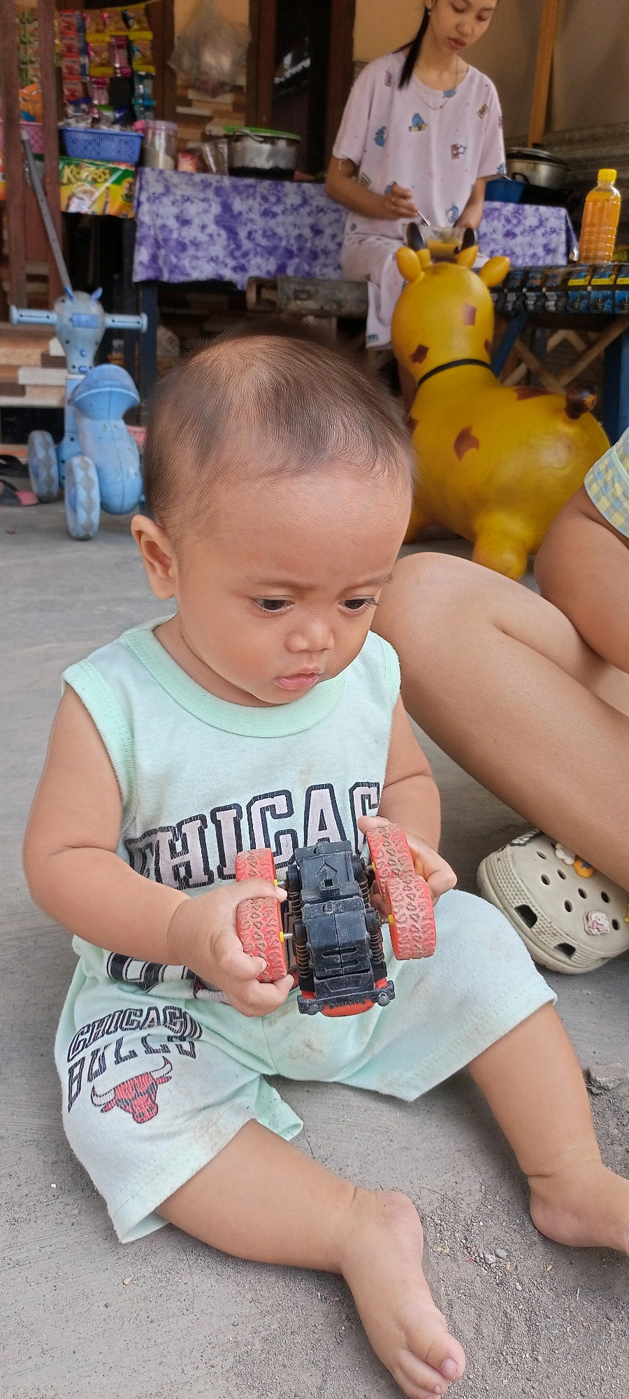 In Gempolklutuk, Indonesia, a young child sits on a concrete surface, holding a black toy car with red wheels. The child wears a light green sleeveless shirt with "CHICAGO" printed on it and light green shorts with a Chicago Bulls logo. To the child's right, a person's legs are visible, wearing a tan Croc shoe. In the background, a yellow toy giraffe is present. Behind the giraffe, a woman in a light pink patterned shirt sits. Further in the background, shelves stocked with various items, including bottled beverages and packaged goods, are visible. A blue toy vehicle with wheels is also located in the background. The environment appears to be outdoors, possibly in front of a shop or market stall.