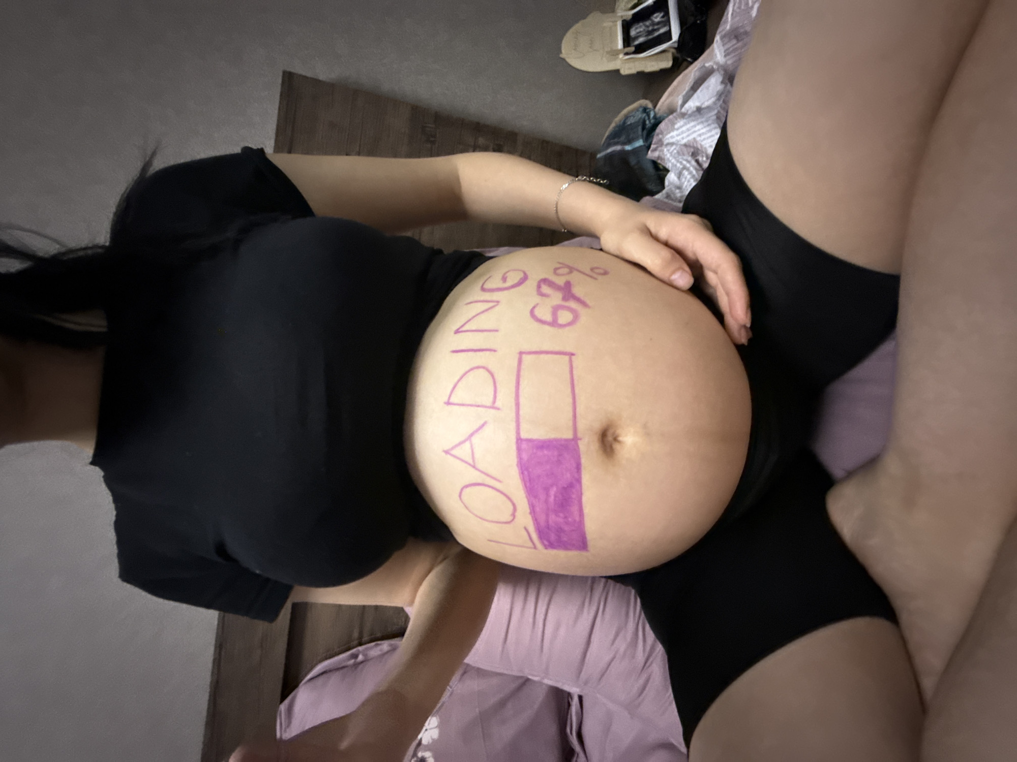A pregnant person's abdomen is decorated with the words "LOADING 67%" written in purple marker. A progress bar is drawn, with the left portion filled in. The person is wearing a black t-shirt and black shorts. They are sitting on a bed with purple pillows. The background shows a wooden headboard and a wall. The scene appears to be indoors, in a bedroom. There is no information to suggest the location is in Tbilisi, Georgia.