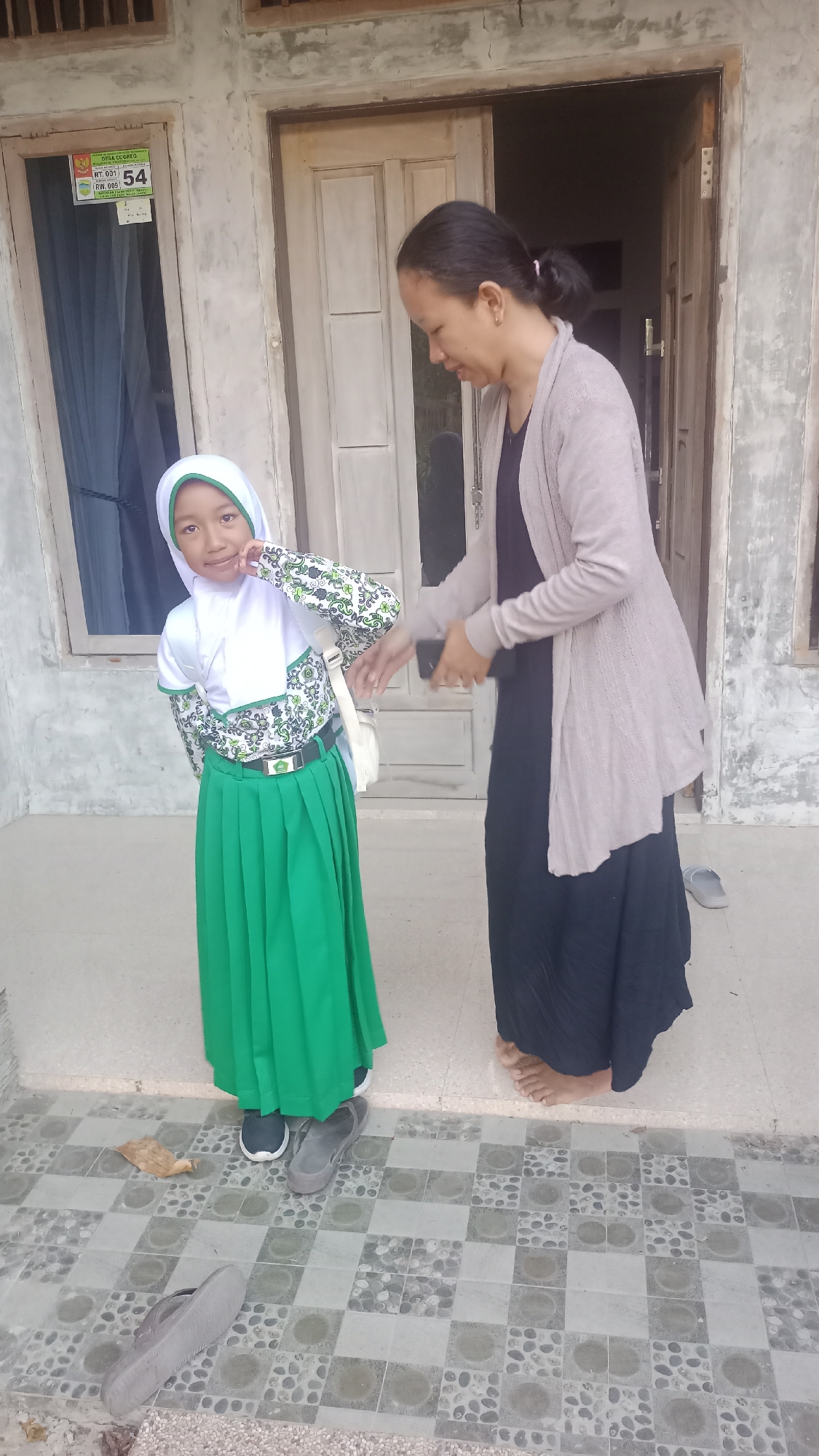 A young girl wearing a white hijab and a green school uniform, complete with a matching green skirt and a white top with green accents, poses for a photo. She has a bright smile and is touching her cheek. A woman, possibly her mother or teacher, stands behind her to the right, wearing a dark dress and a light grey cardigan. She is looking down, possibly at the girl or her phone, which she is holding in her right hand.
They are standing on a tiled porch in front of a building with a light-colored concrete facade and wooden doors. To the left of the girl, a window is visible with a sign attached to it that reads "DESA CIGUBEG RT. 001 RW. 009 54". A pair of grey flip-flops are on the ground near the woman's feet, and another grey shoe is near the girl's feet. A dry leaf lies on the tiled floor. The scene appears to be outdoors, and given the clothing and setting, it is likely a warm day. The image captures a candid moment, possibly before or after school.