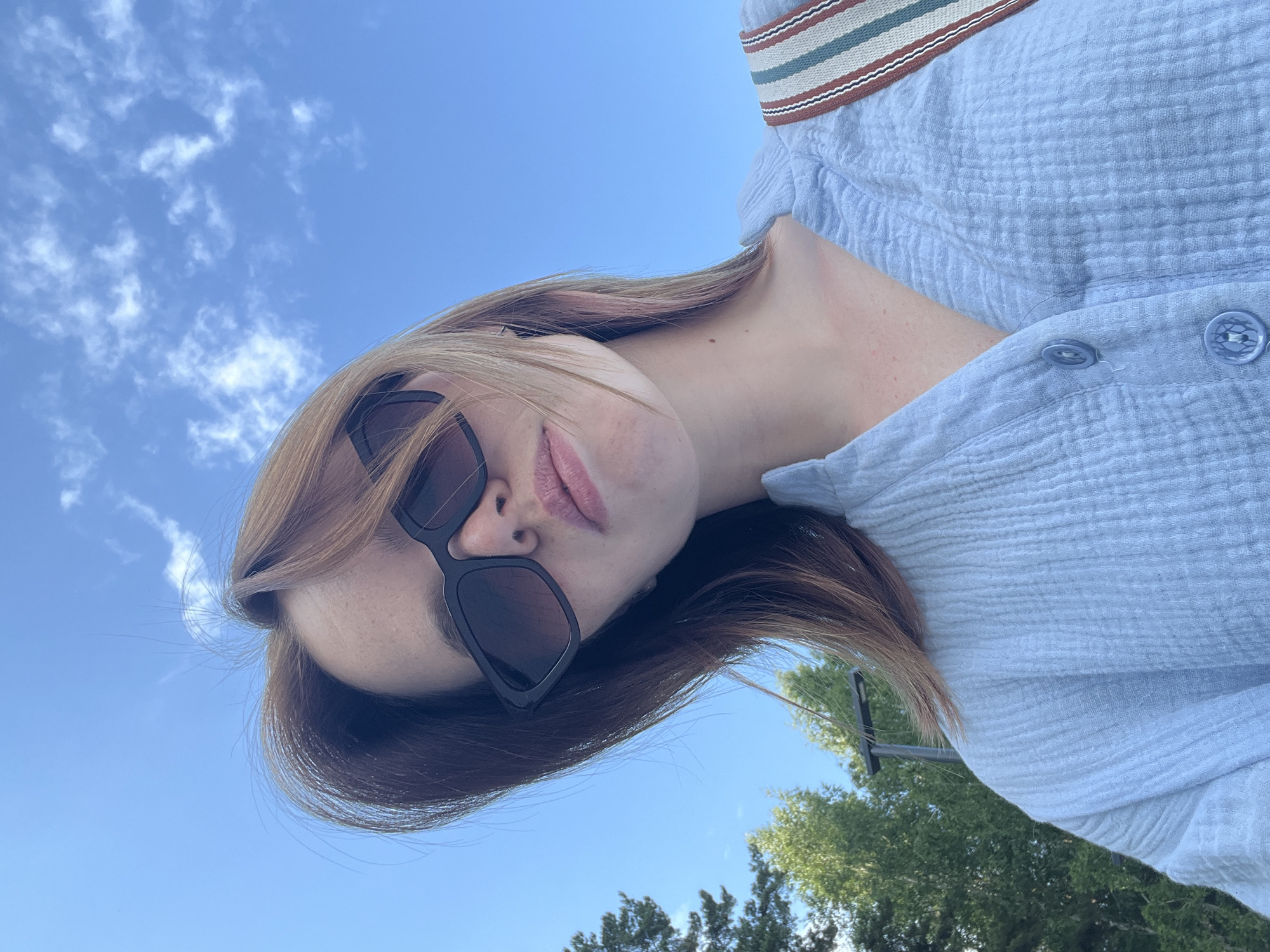 The image is a close-up, low-angle selfie of a young woman outdoors, with a bright blue sky and scattered clouds above. She has medium-length, light brown hair with some lighter streaks and a few pink highlights. She is wearing large, black, angular sunglasses and a light blue, textured, button-up shirt. A portion of a striped bag strap with brown, green, and white stripes is visible over her right shoulder. In the background to the left, several green trees are visible against the sky. The overall impression is of a casual, sunny day. The location is described as Chaikovsky, Russia.