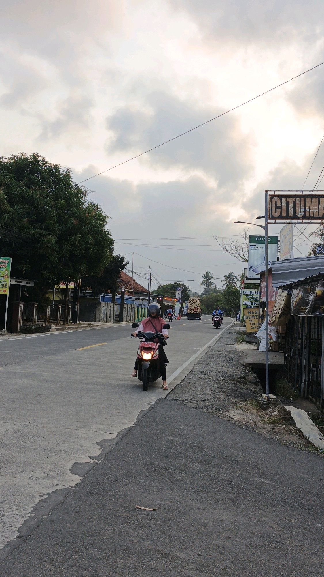This image depicts a roadside scene in Cibenda, Indonesia, under an overcast sky. In the foreground, a woman wearing a pink headscarf and a pink long-sleeved shirt is riding a dark-colored scooter. She is facing forward, looking ahead. The scooter has a license plate that reads "F 2053". The road in front of her is paved, with a white line marking the lane. To the right of the road is a dirt and asphalt shoulder with some sparse vegetation.
Along the right side of the road, there are several structures. A prominent sign with the word "CITUMAN" in large, distressed letters is visible. Below it, there are other signs, including one that says "OPPO". Further along the right, a stall-like structure with a dark corrugated metal roof is present. Various goods are displayed on shelves outside this stall, including bags of items and some packaged goods.
On the left side of the road, there are trees, fences, and buildings. A large tree with dense green foliage dominates the left side of the image. Behind the tree, there are houses with reddish-brown roofs and fences.
In the background, the road continues, with other vehicles visible. A truck with a large load of logs is driving away from the camera. Other motorcycles are also present on the road. Palm trees and other vegetation can be seen in the distance, suggesting a rural or semi-rural environment within Cibenda. The overall atmosphere is calm, with the muted light indicating it might be early morning or late afternoon.