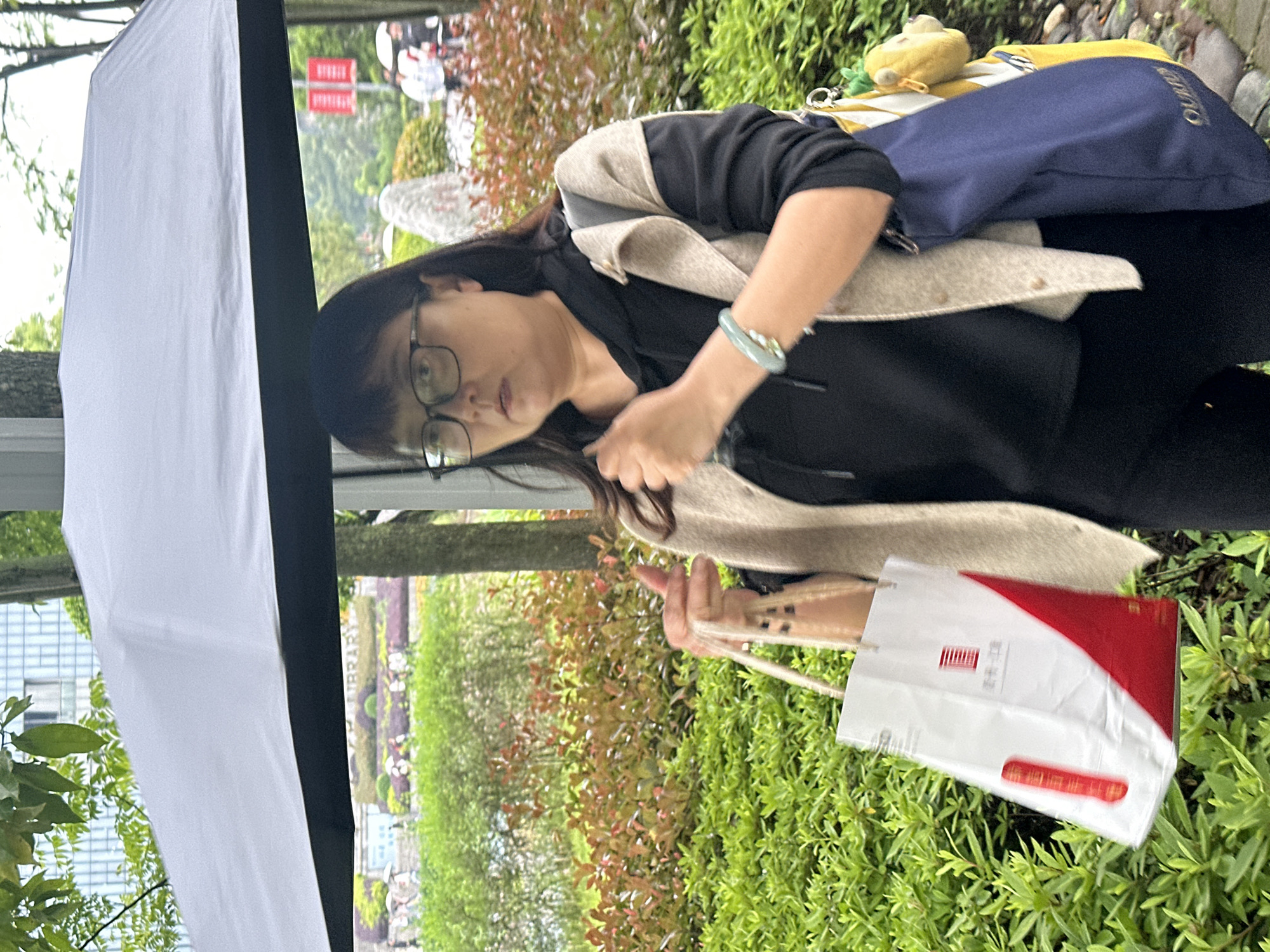 A woman with glasses, wearing a black top, beige cardigan, and a blue jade bracelet, is holding a shopping bag with a red and white design. She also has a blue bag with a yellow and white checkered section and a yellow plush charm on her shoulder. She appears to be standing outdoors near shrubbery, a tree, and a large white fabric surface. In the background, a structure with the word "LIBRARY" visible is identifiable as the Jiangjin Library in Jiangjin, China.