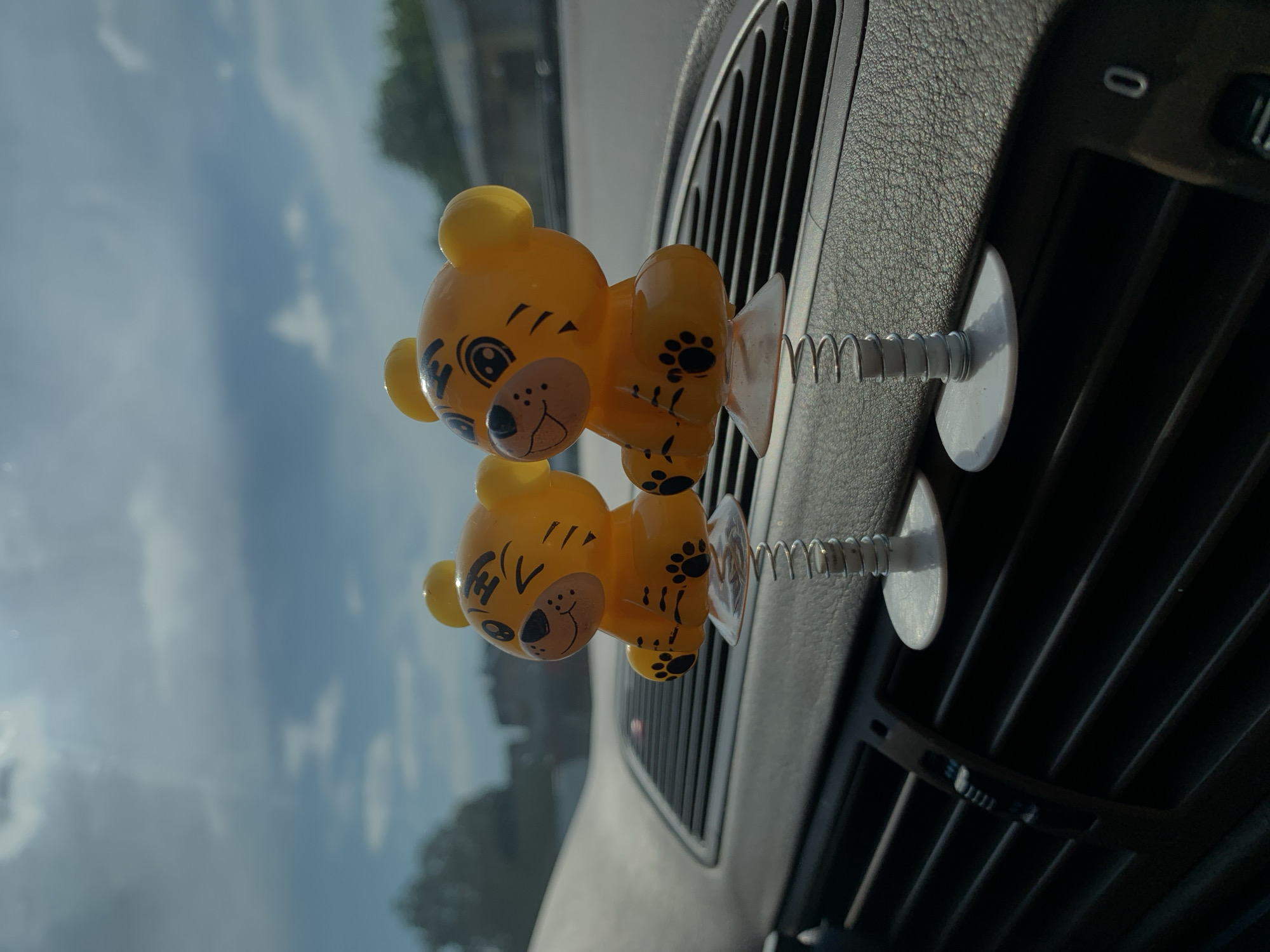 The image displays a close-up, eye-level perspective of two yellow, plastic, cartoon-style tiger figures positioned on the dashboard of a vehicle in Harmanli, Bulgaria. Each figure is affixed to a coiled metal spring, which is attached to a clear plastic suction cup adhered to the textured, dark grey dashboard surface, directly above a car air vent.
The left figure exhibits a winking left eye and a smiling expression, with painted black details including an oval nose, mouth, three facial dots, stylized stripes on its forehead and cheeks, and distinct paw prints on its abdomen and feet. The right figure, positioned slightly behind and to the right, shows open eyes and a broader smile, with similar black painted features and paw prints. Both figures have rounded ear nubs.
The dashboard features a visible granular texture, and the air vent beneath the figures has horizontal slats. Through the vehicle's windshield, an overcast sky with patches of blue is visible, along with blurred green foliage from trees and indistinct distant structures. The scene is illuminated by natural daylight. No people are present.