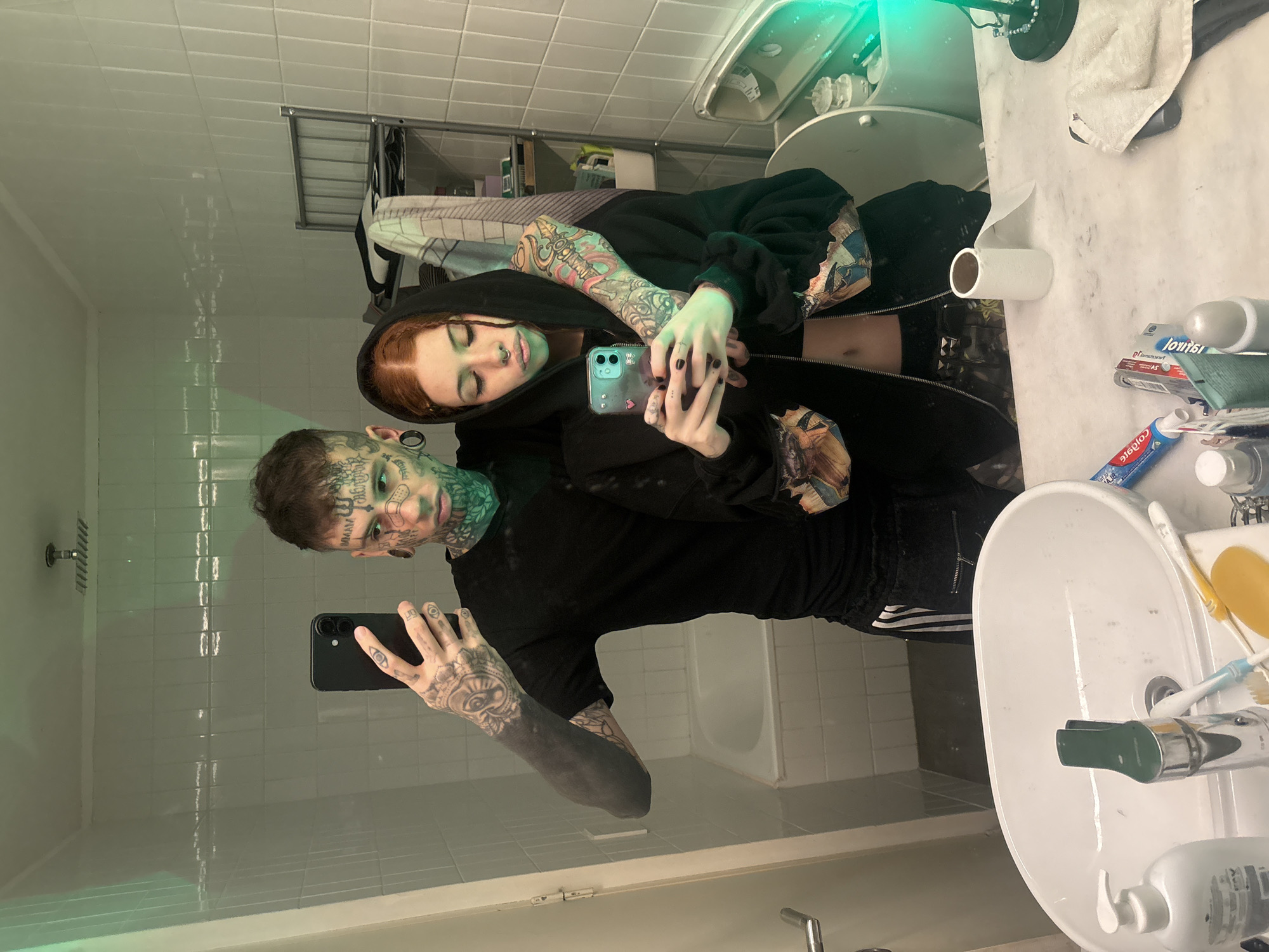 A man and woman are taking a selfie in a bathroom mirror. The man on the left is holding a black smartphone. He has numerous tattoos on his face, neck, and arms. The woman on the right, wearing a black hoodie, holds a blue smartphone. She also has tattoos on her arms. The background reveals a standard bathroom setup: white tiles, a bathtub, a sink with toiletries, and a toilet. The location is not discernible from the image.