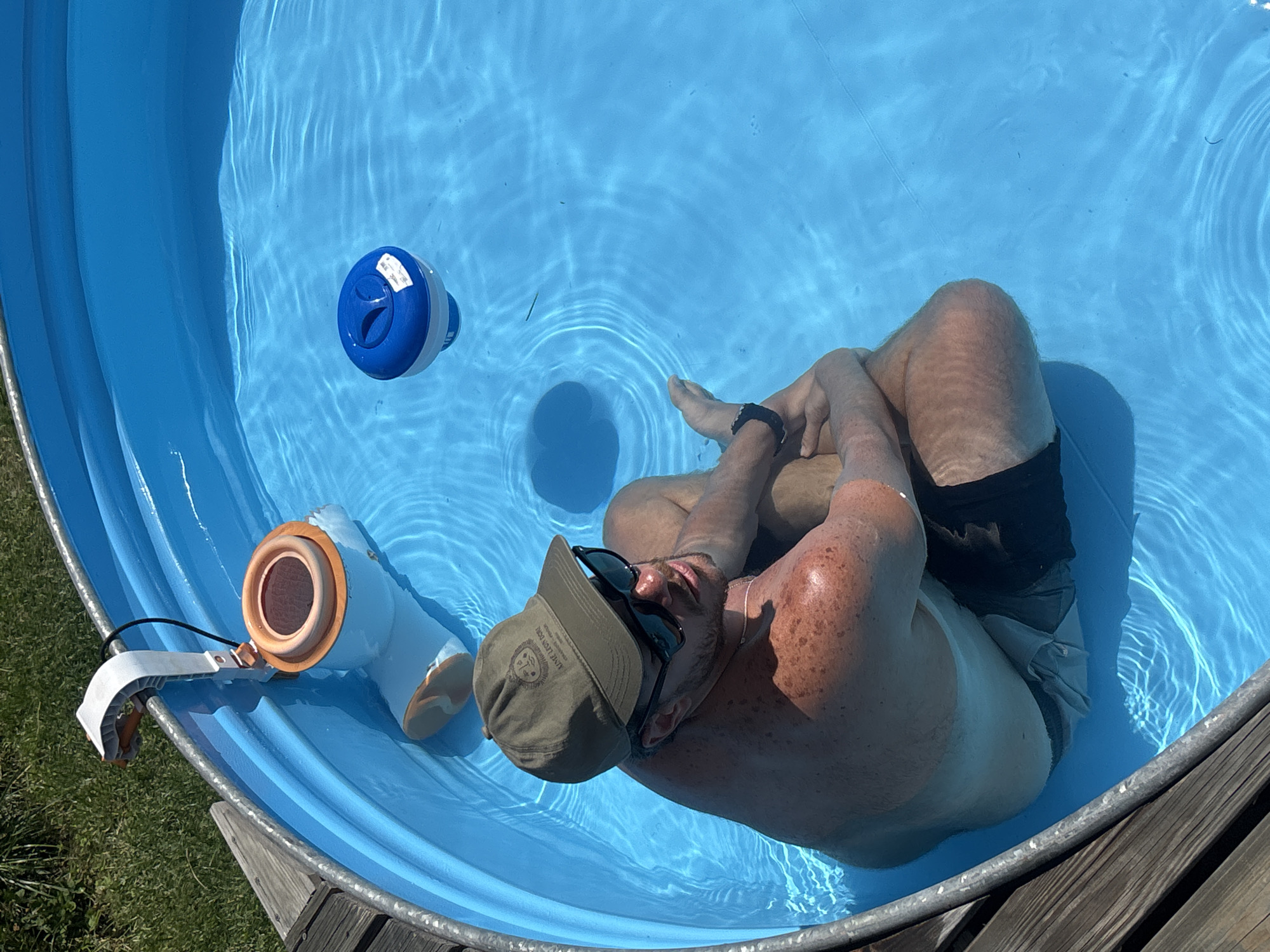 A medium shot from above shows a man sitting in a circular, above-ground swimming pool filled with blue water. He is wearing a green baseball cap, sunglasses, and dark swim trunks. His arms are crossed, and he appears relaxed. The pool has a metal rim and a wooden deck surrounding it. In the background, there is green grass and a pool skimmer attached to the rim, with a blue chemical dispenser floating in the water. The sun is shining, casting shadows on the water. The image's metadata indicates the location as San Lorenzo de Tormes, Spain.
