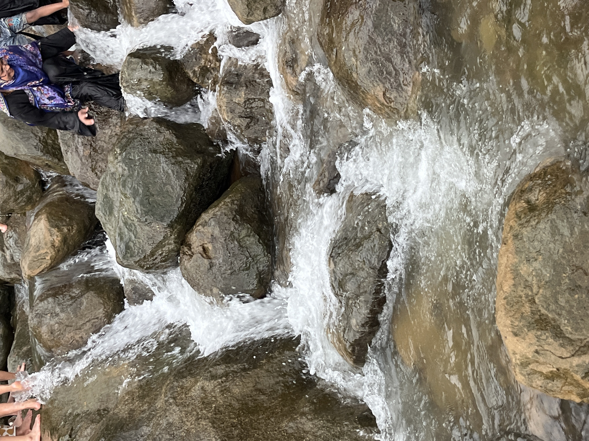 In Jaflong, Bangladesh, the image captures a close-up view of a shallow river with clear, flowing water cascading over numerous rocks. Several people are present, some wading in the water with their feet visible, while others, including a woman in traditional attire, are navigating the rocky terrain near the water's edge. The environment is characterized by a rocky riverbed and surrounding boulders, with the water appearing somewhat murky. The scene depicts a natural setting with human interaction, suggesting activities such as crossing the river or enjoying the water.