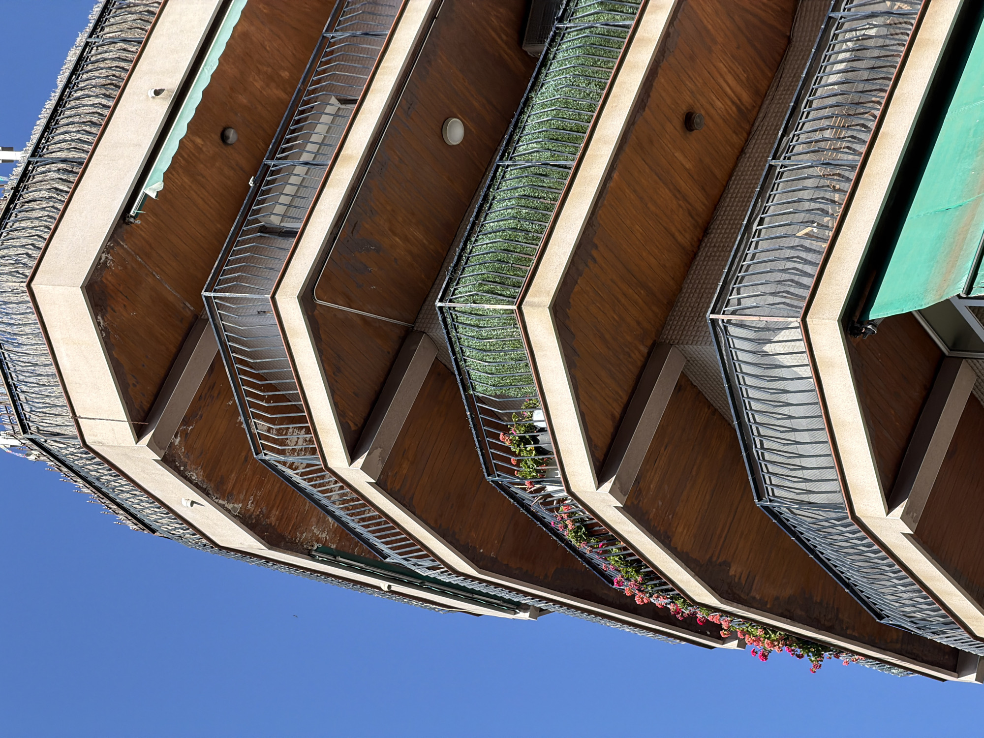 This is a low-angle, upward shot of the underside of multiple balconies of a modern apartment building, against a clear blue sky. The balconies have a distinctive zigzagging, angular design, with wooden soffits and light-colored structural beams. Each balcony is equipped with a metal railing and some contain planters with blooming pink and red flowers, as well as some artificial green foliage. Some balconies also feature awnings. The scene suggests a sunny day, likely in the late morning or early afternoon given the bright light and deep shadows. The architecture is contemporary and could be found in a Mediterranean city, with Barcelona being a likely candidate. There are no people visible in the frame, and no text is present. The overall impression is one of vibrant colors against a minimalist architectural backdrop.