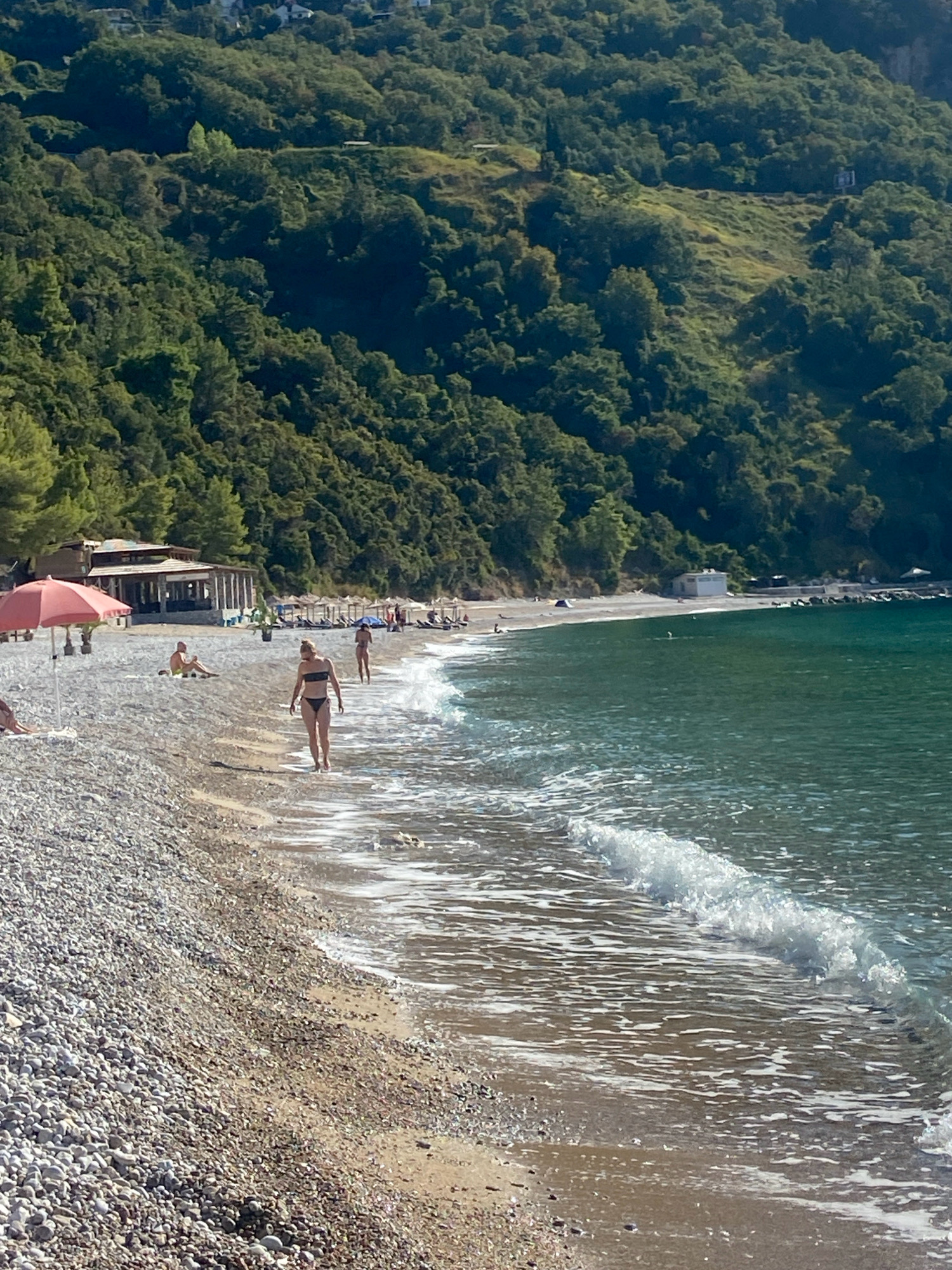 This image depicts a pebble beach with a calm, turquoise sea in the city of Prijevor, Montenegro. In the foreground, the water gently laps at the shore, creating white foam. A woman in a black bikini walks towards the water, with another woman in a bikini further down the beach, walking away from the camera. To the left, a pink umbrella shades a beach chair, and a person is seated on the sand. In the midground, a row of sun loungers with umbrellas is set up along the beach, leading to a covered structure that appears to be a restaurant or bar. The background is dominated by a densely wooded hillside that rises steeply from the coastline. The sky is clear and blue, suggesting a sunny day.