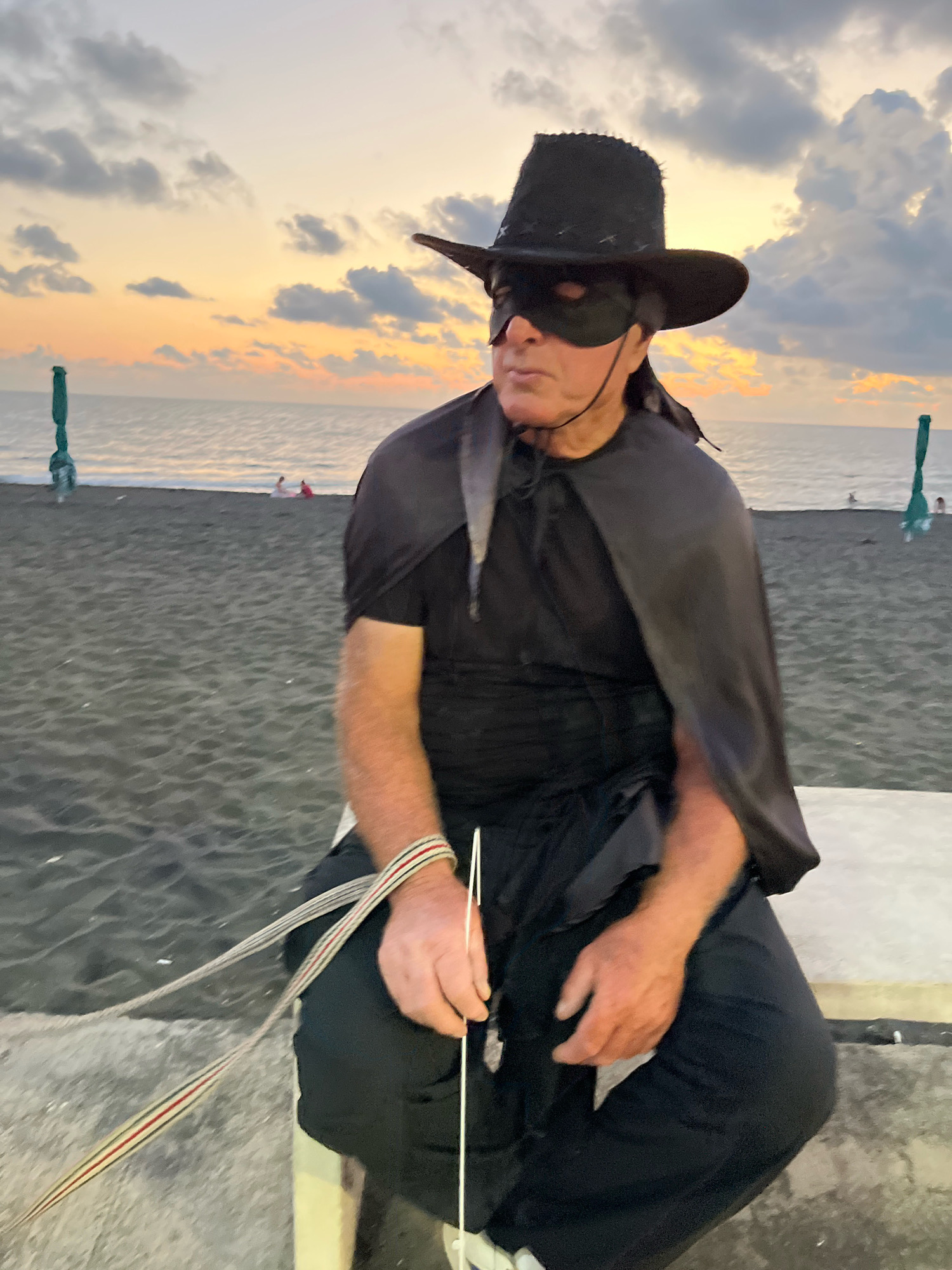 A man dressed as Zorro sits on a low concrete wall on a black sand beach in Ureki, Georgia. He wears a black wide-brimmed hat, a black eye mask, a black cape, and black pants. His left hand rests on his lap, holding a white object with a thin cord attached. A striped ribbon is wrapped around his left wrist. In the background, the sea meets the horizon under a sky filled with soft, colorful clouds, indicative of sunset or sunrise. Two green beach umbrellas are visible on the beach.
