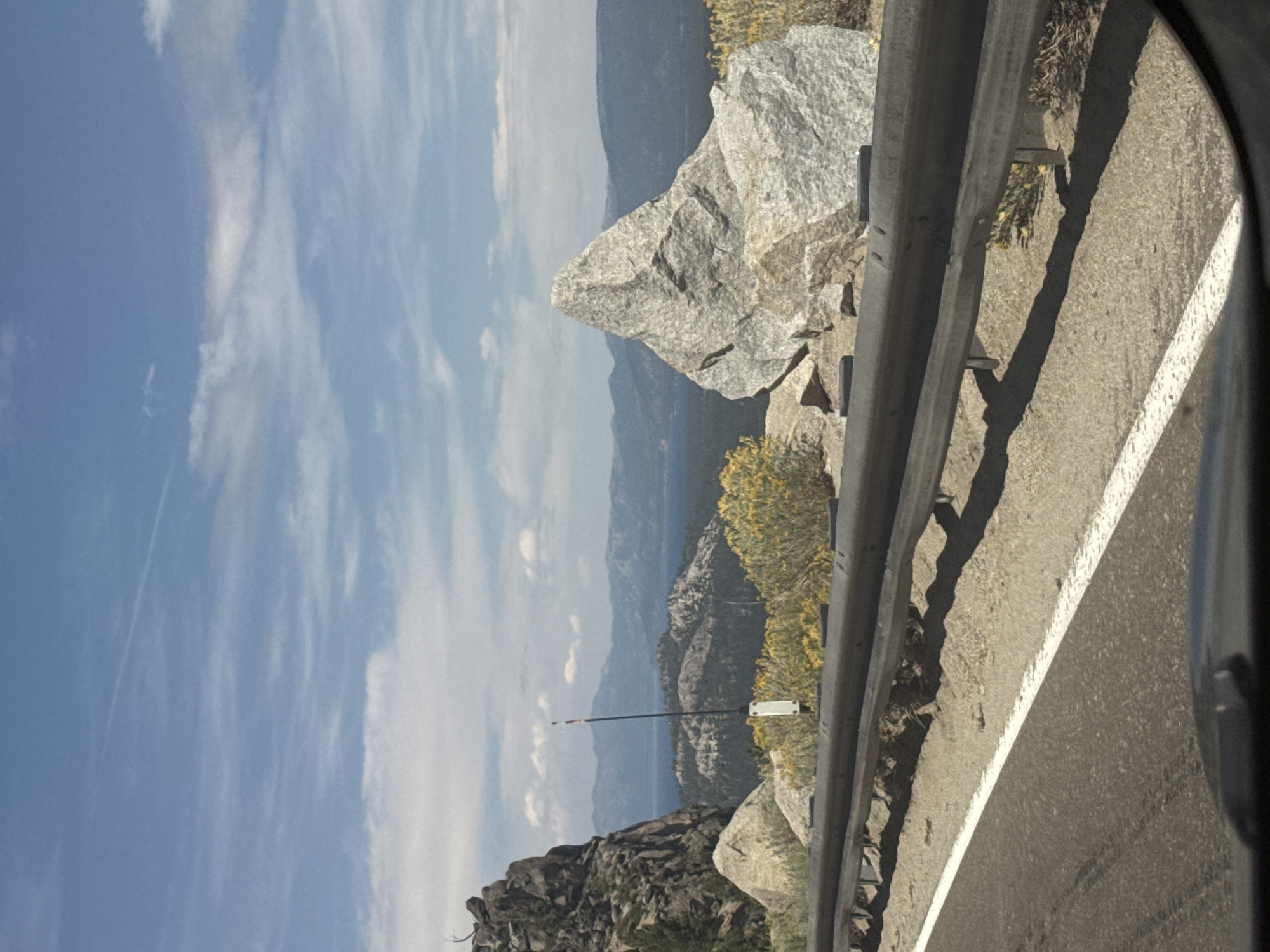 The image shows a scenic overlook on a sunny day, with a view of a large lake and mountains in the distance. The foreground features a guardrail along a paved road. To the left, a large, dark rock formation juts out, partially covered in greenery. To the right of the rock formation, a tall, thin pole with a white base and a small, multi-colored flag or antenna at the top stands prominently. In the mid-ground, a large, light-colored boulder with a pointed top is situated, with sparse yellow-green vegetation growing around its base and behind it. The background reveals a vast expanse of blue lake water, bordered by rolling, tree-covered hills and distant mountain ranges under a bright blue sky with scattered wispy white clouds. The overall impression is of a natural, mountainous landscape with a developed roadside.