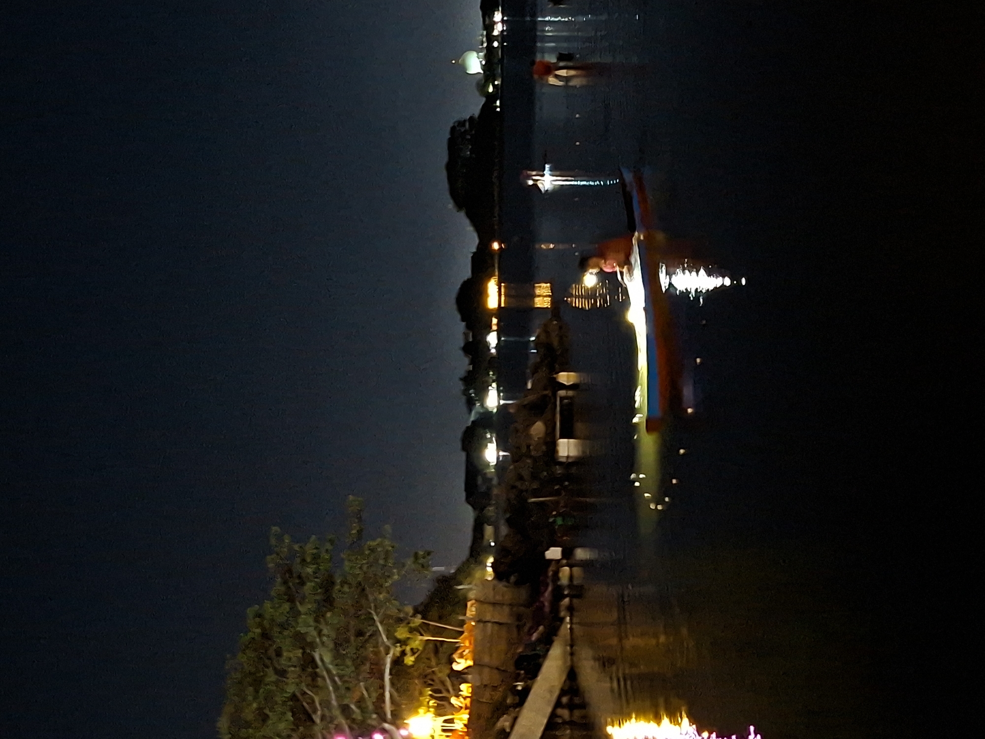 This is a nighttime photograph of a body of water, likely a bay or river, in Lhokseumawe, Indonesia. The scene is illuminated by artificial lights, creating reflections on the dark water. In the foreground, a person is sitting in a small boat, holding a lantern that casts a warm glow. The boat appears to be a kayak or a similar small watercraft. Further in the distance, other figures are visible on the water, some of whom are also illuminated, suggesting they might be participating in a similar activity or are in other boats.
On the shore to the left, trees are visible with decorative lights. In the background, there are buildings and structures, some of which are lit, indicating a populated area. A prominent dome, possibly belonging to a mosque or another significant building, is visible in the far background to the right, silhouetted against the night sky. The overall atmosphere is serene and festive, with the lights creating a magical ambiance on the water. The water itself is calm, with gentle ripples reflecting the light sources. The sky is dark and clear, characteristic of a night scene in a tropical location. The image captures a moment of activity and community on the water at night.