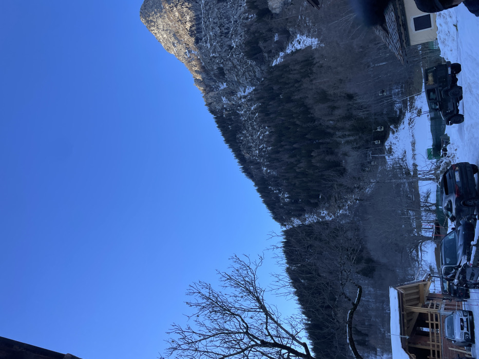 This image, originally captured in portrait orientation and rotated 90 degrees counter-clockwise, depicts a bright winter scene in Courmayeur, Italy. The vast, clear blue sky dominates the upper left portion of the frame, indicating good weather.
A large, rugged mountain, partially covered in snow and rock, rises prominently on the right, bathed in strong sunlight. Below the mountain, a dense evergreen forest blankets the slopes, also showing signs of snow.
In the midground and foreground, on the bottom right, is a snowy parking area occupied by several vehicles, including a large, dark SUV resembling a Hummer H3 and other passenger cars. To the far right, a building with a dark roof and light walls is visible, possibly part of a resort. Closer to the foreground, a wooden structure with a snow-covered roof and exposed beams appears to be under construction or recently built. A prominent bare-branched deciduous tree stands in the lower right, adding to the winter aesthetic. Faint structures resembling ski lift components can be discerned among the trees in the midground.
The scene is set during daylight, likely morning or early afternoon, given the clear sky and bright illumination on the mountain and foreground elements. The presence of snow, ski lift elements, and resort-style buildings suggests a winter sports destination. No people or visible text are discernible in the image.