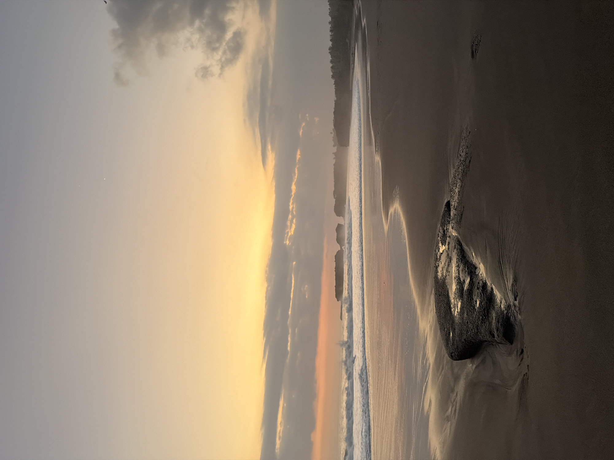 This vertical image captures a coastal landscape at twilight in Selemadeg Timur, Indonesia. The composition is divided into three primary horizontal sections: sky, ocean and distant land, and foreground beach.
The upper half of the frame displays a sky gradient, transitioning from a luminous yellow-orange at the horizon to a muted grey-blue overhead. Scattered cloud formations are present, some illuminated with warm tones near the horizon, while others appear darker and denser in the upper right. Two small, distinct bright points of light are visible in the upper-left sky. A dark, indistinct shape, likely avian, is present in the far upper-right corner.
The middle ground features the ocean, with white-capped waves actively breaking onto the shore. In the mid-distance, several dark, rugged rock formations or small islands emerge from the water. To the right, a coastline curves, revealing a dense band of dark green tropical foliage, consistent with a treeline, extending along the shore. Haze or mist partially obscures the distant landforms.
The foreground is composed of a dark, fine-grained sandy beach, exhibiting patterns and textures from receding tides. The wet sand near the waterline reflects the warm light of the sky. A single, prominent, irregularly shaped dark rock, partially embedded in the sand and wet from the ocean, occupies the lower-left quadrant of the beach. No human figures or discernible animals are present in the scene. The lighting indicates either sunrise or sunset conditions.