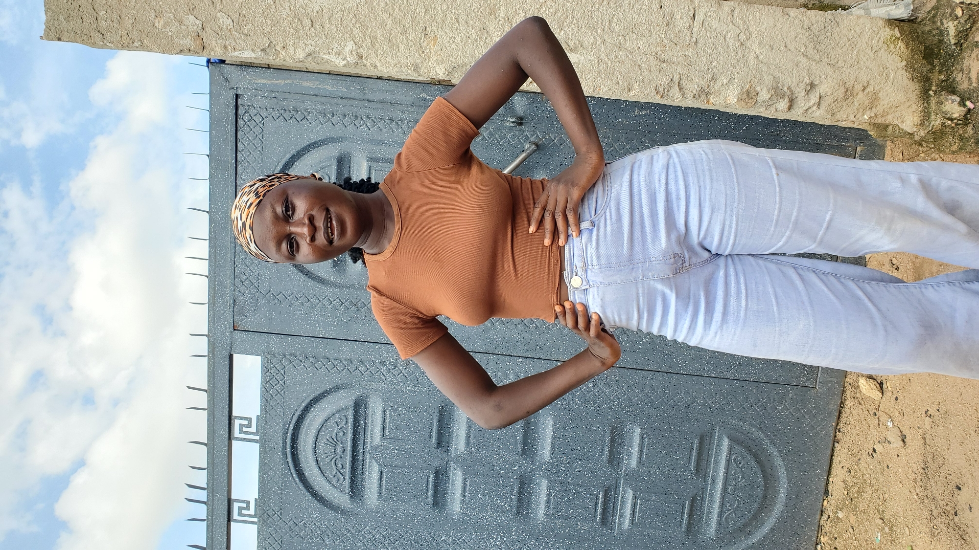 A young Black woman stands in front of a grey, ornate metal gate in Jalingo, Nigeria. She is wearing a brown, ribbed crop top and light blue, wide-leg jeans. A patterned headscarf adorns her head. Her hands are on her hips, and she is looking at the camera with a slight smile. The gate is closed and has a decorative pattern of circles and Greek key designs. To the right of the gate is a rough, grey concrete pillar. The ground in front of the gate is sandy and strewn with small stones and debris. The sky above is blue with scattered white clouds.