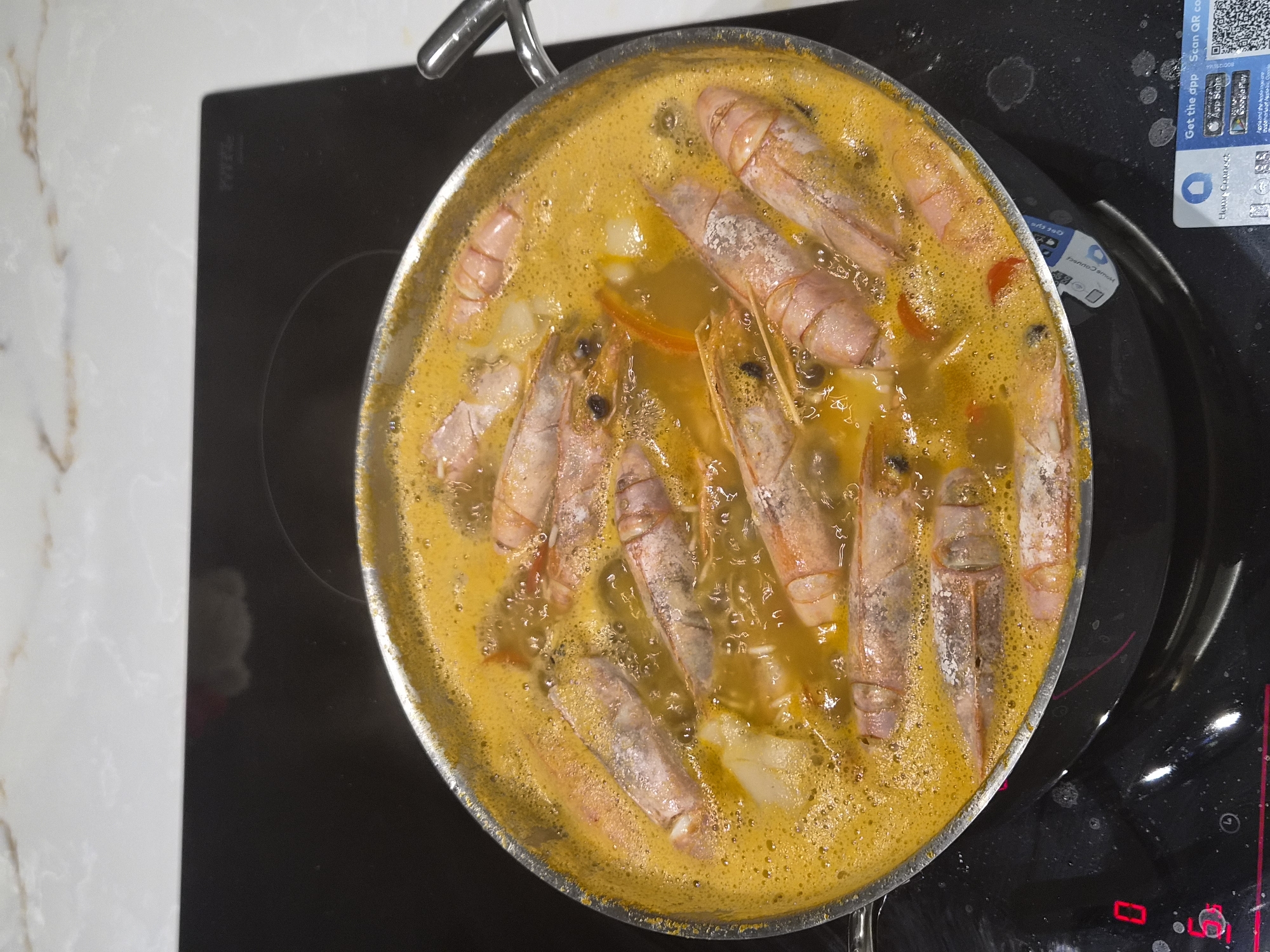 A close-up, overhead shot shows a metal pot filled with a bubbling, orange-yellow broth containing several large, cooked prawns and what appear to be pieces of white onion and red pepper. The pot is placed on a black induction stovetop. The stovetop displays a red "0" on the left and a partial "5" with a circle around it below. To the right of the pot, a small QR code sticker is visible on the stovetop, with text indicating "Get the app. Scan QR code." The background consists of a marble countertop with light beige and white veining. The image does not contain any identifiable landmarks or specific details that would indicate its location within the city of Barcelos, Portugal.
