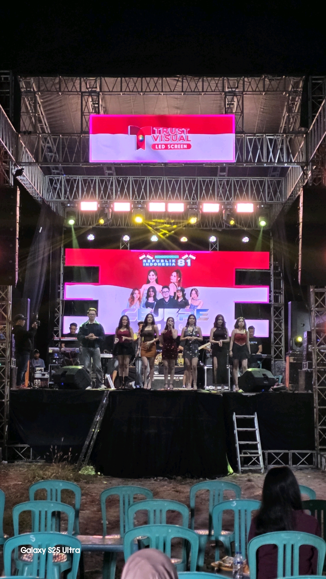 A stage with a large LED screen is set up outdoors at night in Jaken, Indonesia. The screen displays a red and white graphic with text that reads "REPUBLIK INDONESIA 81" and images of several women and one man. A group of women in dresses are standing and singing on the stage in front of the screen. A man with glasses is standing and speaking to the audience on the stage. Another man is standing on the left side of the stage holding a camera. There are several musicians in the background on the stage playing instruments. In the foreground, there are rows of teal plastic chairs facing the stage, some of which are occupied by people. A ladder leans against the side of the stage. The ground appears to be dirt. The overall scene suggests an outdoor concert or performance.
