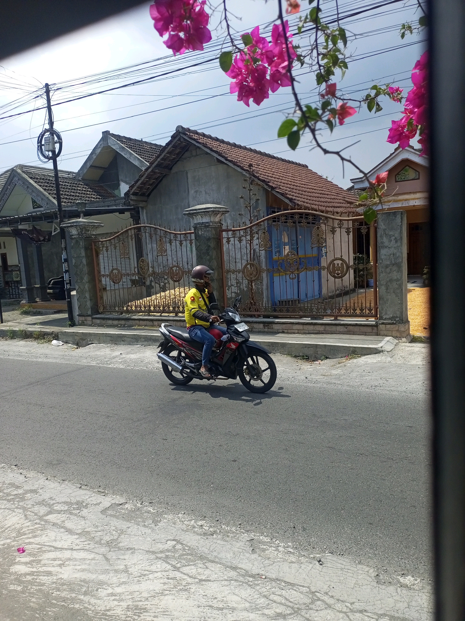A person wearing a yellow long-sleeved shirt, blue jeans, a maroon helmet, and white sandals rides a red and black motorcycle down an asphalt road. The motorcycle has a silver exhaust pipe. The person is holding a black mobile phone to their right ear with their left hand. In the background, there is a fence made of ornate metal bars and concrete pillars. Behind the fence are several houses with tiled roofs and grey walls. One house has a blue metal door. Pink flowers are visible in the foreground, hanging from branches. The sky is cloudy. The location is identified as Ngasem, Indonesia.