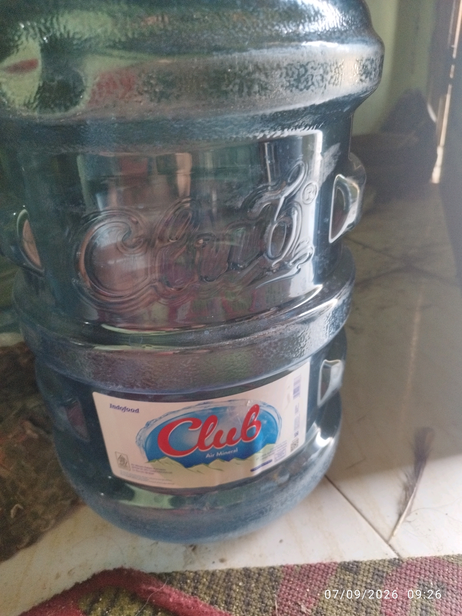 A close-up, eye-level shot shows the bottom section of a large, clear plastic water jug, likely filled with water, resting on a tiled floor. The jug has a textured band around its middle and the top of the jug is visible. A label is affixed to the lower part of the jug, featuring the brand name "Club" in red and blue lettering, with "Air Mineral" below. The text "Indofood" is also visible on the label. The floor appears to be light-colored tile. In the foreground, the edge of a textured mat or rug is visible, with a timestamp in the lower right corner reading "07/09/2026 09:26". The lighting suggests an indoor environment, possibly with natural light filtering in from the right side.