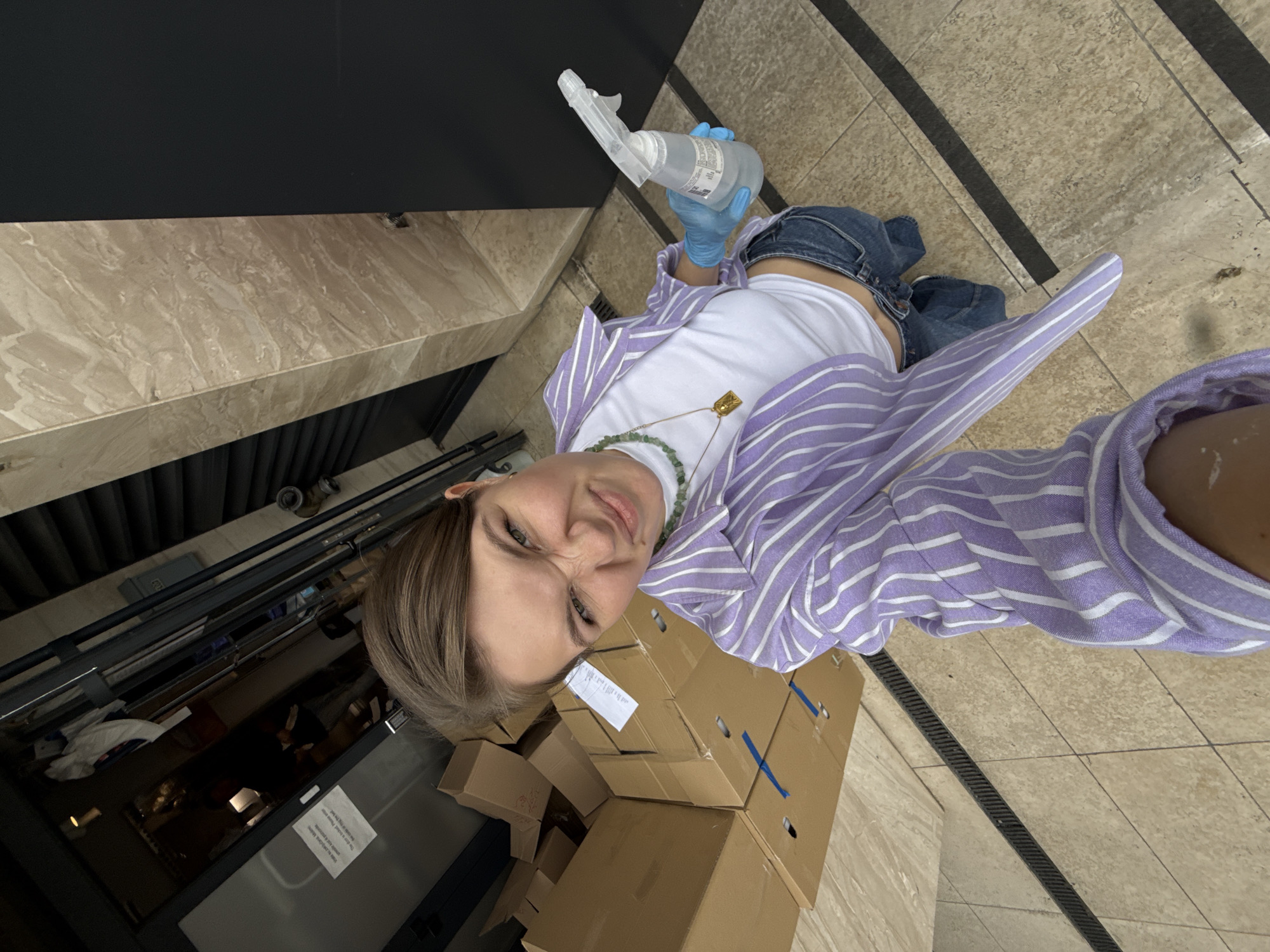 A young woman is seen indoors, possibly in Belgrade, Serbia. She is holding a spray bottle with a clear liquid and wearing blue gloves. She has on a white top, a lavender striped shirt, and blue jeans. She is wearing a necklace with a gold pendant and a green beaded necklace. Beside her is a stack of cardboard boxes. To the left is a glass-fronted cabinet or refrigerated display case. The floor is tiled.