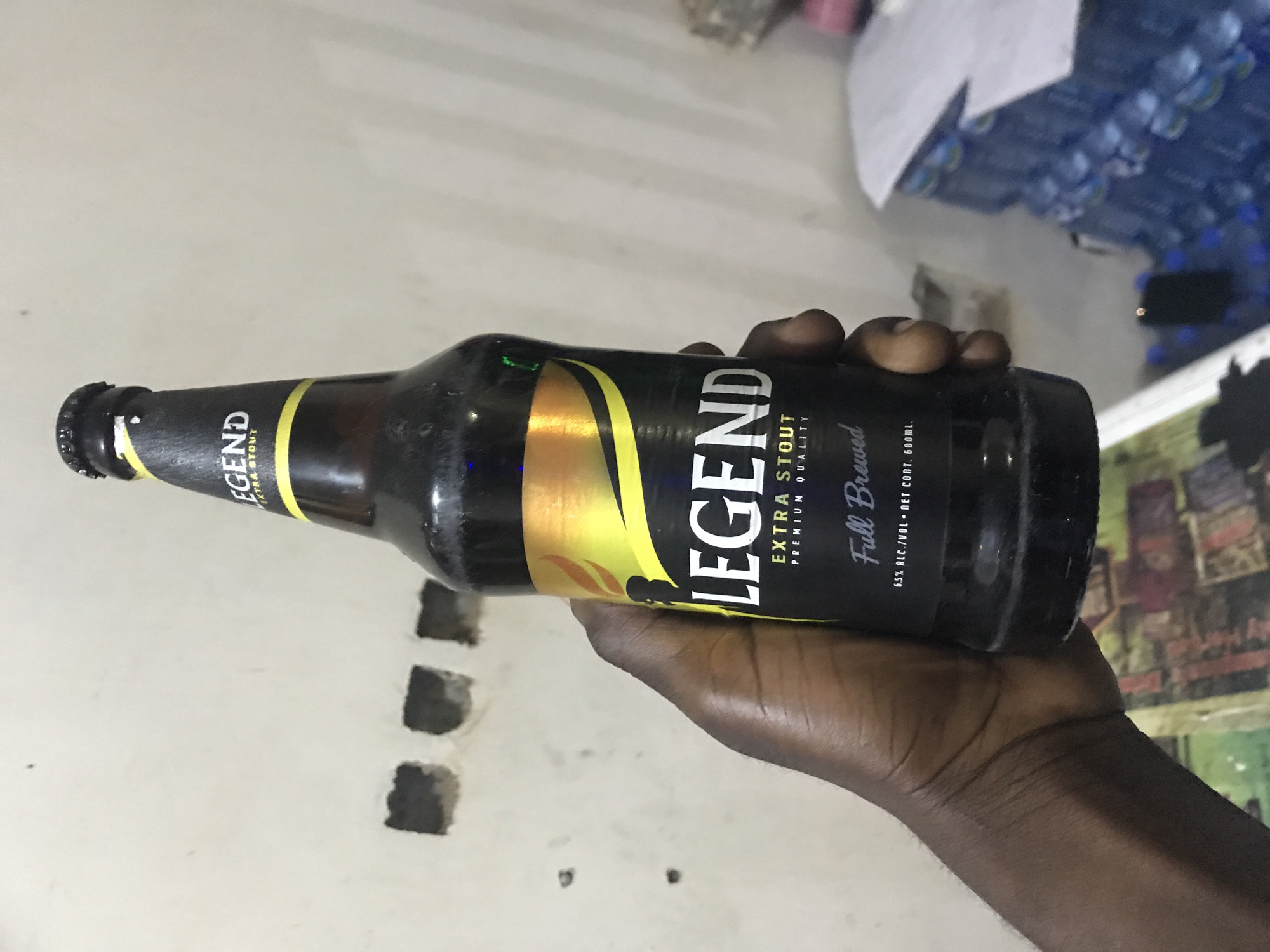 A dark-skinned hand holds a brown bottle of "Legend Extra Stout Premium Quality Full Brewed" in an indoor setting. The label has a yellow and gold design element. The text on the label indicates the alcohol content is 6.5% by volume and the net content is 600 ml. The wall adjacent to the hand is white with several dark, irregularly shaped holes. The bottle is held horizontally. A collection of clear plastic water bottles is visible in the background, along with a piece of paper and a dark electronic device. The image was likely taken in Kabong, Nigeria, as specified in the prompt.