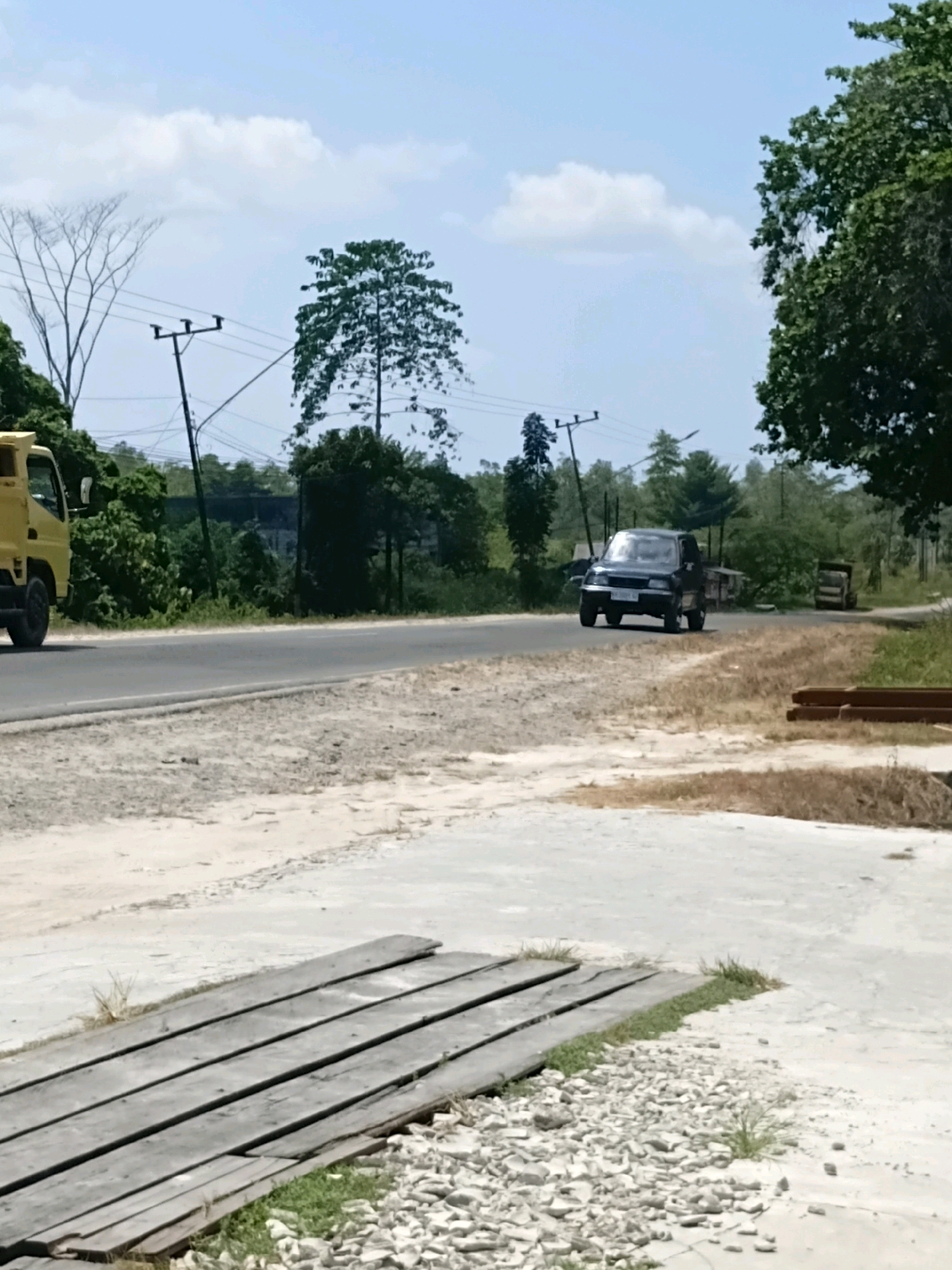 The image depicts a roadside scene in Batu Belaman, Indonesia. A paved road runs horizontally across the frame, bordered by a gravelly shoulder. On the left side of the road, a yellow truck is partially visible, moving away from the viewer. Further down the road, a dark-colored passenger car is approaching the camera. The background is filled with lush green trees and vegetation, with utility poles and power lines visible overhead. The sky is bright blue with scattered white clouds. In the foreground, wooden planks are laid out on the ground, interspersed with small stones and patches of grass.