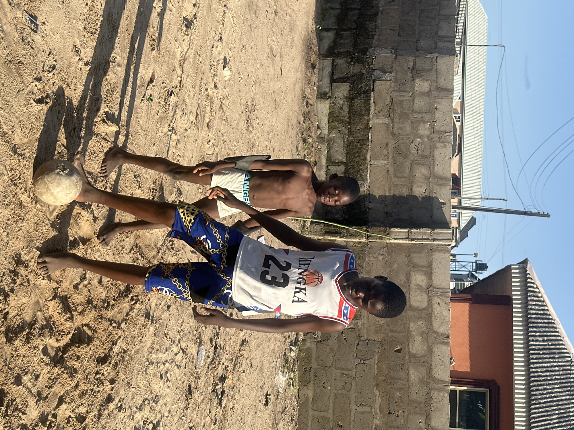 Two young boys are in a sandy outdoor area, likely a street or yard, in Badagry, Nigeria. The boy in the foreground is wearing a white basketball jersey with the number 23 and the word "MENGKA" printed on it, along with blue and gold patterned shorts. He is holding a soccer ball with his left hand and looking towards the viewer. The second boy, slightly behind and to the left, is wearing white shorts. Both boys appear to be of African descent and are shirtless.
The scene is set during the daytime, with strong shadows cast on the sand, suggesting bright sunlight. The background features a brick wall and parts of buildings, some with corrugated metal roofing, and power lines overhead. The overall setting suggests a warm, possibly hot, climate. The boys' expressions are neutral, and the activity appears to be playing or posing with a soccer ball. The lighting and shadows indicate it could be late morning or early afternoon.
