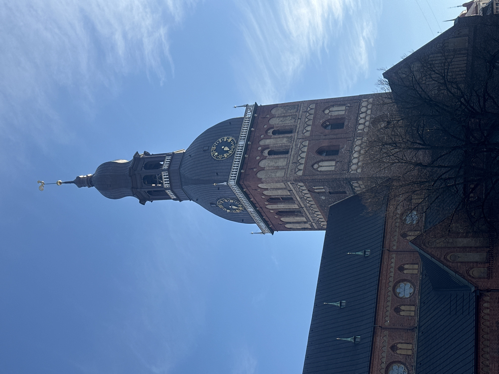 The image shows the tower of St. Peter's Church in Riga, Latvia, viewed from below against a blue sky with wispy clouds. The tower is made of red brick, with arched windows and decorative elements. The top of the tower features a clock and a dark-colored spire topped with a golden weather vane. Bare tree branches are visible on the right side of the frame. A section of the church's roof is also visible, covered with dark tiles and several spire-like decorations.