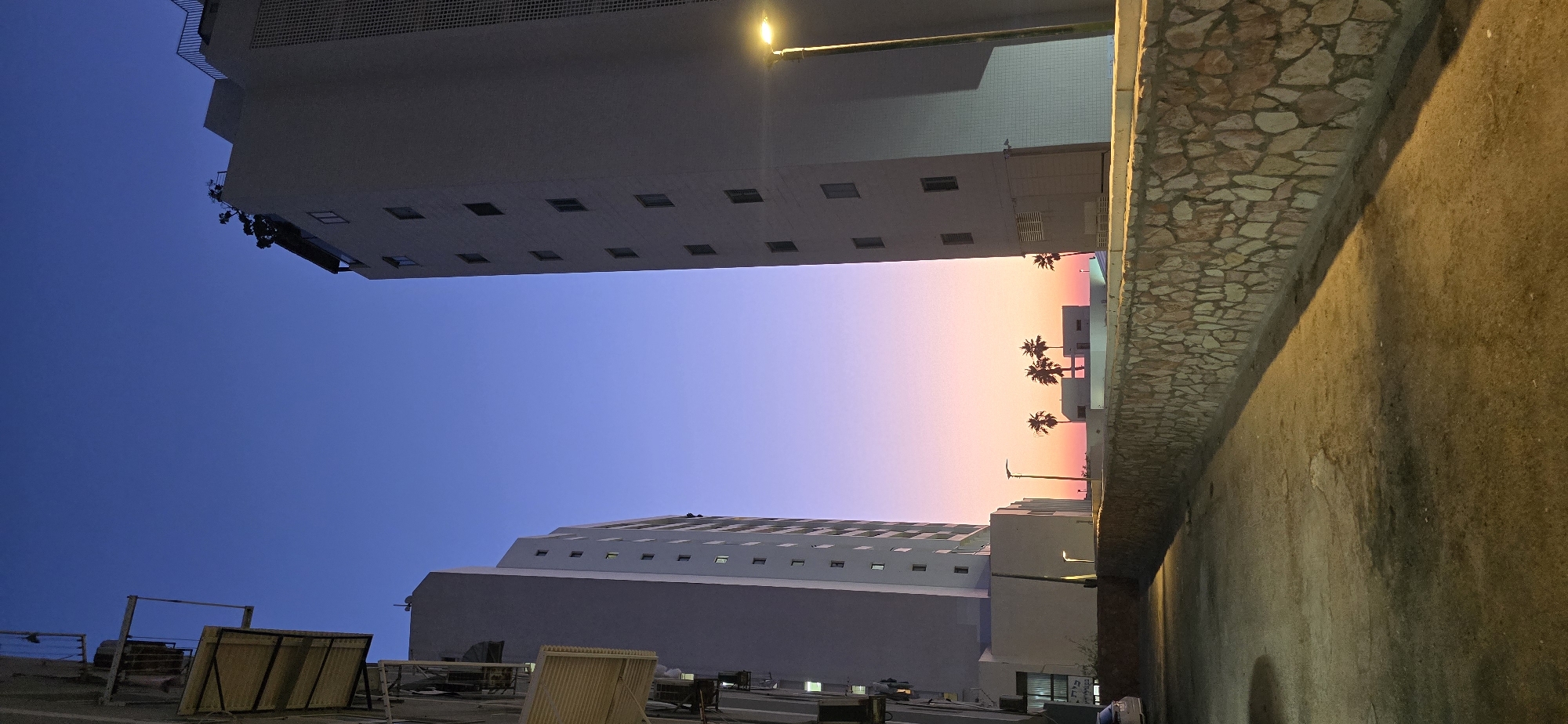 This is a full-frame, vertical, eye-level shot of apartment buildings in Bat Yam, Israel, at twilight. The sky displays a gradient of colors, transitioning from deep blue at the top to pink and orange near the horizon. Two tall, light-colored apartment buildings dominate the frame, one on the left and one on the right, creating a canyon-like effect. The building on the left has multiple balconies with awnings. The building on the right features a textured facade and a prominent streetlamp emitting a warm yellow light. In the distance, between the buildings and at a lower elevation, are smaller white structures and several palm trees silhouetted against the colorful sky. A low, rough-hewn stone wall runs along the bottom foreground, with a paved area in front of it. The overall scene suggests a coastal urban environment.