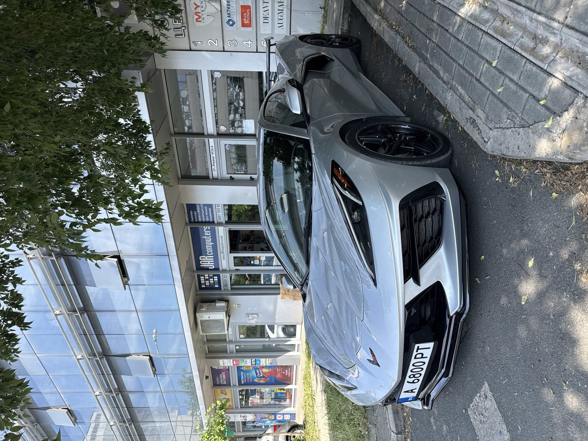 A silver-grey Chevrolet Corvette C8 sports car is parked on the side of a street in Burgas, Bulgaria, during the daytime under clear skies. The car, displaying a Bulgarian license plate "A 6800 PT" on its front bumper, faces towards the left, showcasing its sleek design, black multi-spoke wheels, and dark front grille.
To the left of the car, a modern building with a reflective glass facade and several air conditioning units is visible. A large green tree partially covers the upper left portion of the building and the sky. The building houses various businesses, identified by multiple signs.
Visible text on the building signs includes "JAR computers" with its website "www.JARComputers.com" and phone numbers "056 531 222", "0887 955 85". Other listed services are "Компютри" (Computers), "Аксесоари" (Accessories), "Мрежи и Модули" (Networks and Modules), "Компютърни услуги" (Computer services), and "Консумативи" (Consumables). Additional business names like "MY INTERN", "АКТИВИ КОНСУЛТ", "plusminus", "DEAVIT", and "AUGMAF" are displayed alongside numbers 1, 2, 3, 4. A street sign reads "ул. „Вардар" №26 A" (Vardar Street No. 26 A). Other texts visible are "ГРИЖА ЗА СТАВИТЕ" (Care for joints), "МАКСИМАЛНО НОЖЕСТВО", and "COLOREX".
The car is parked on asphalt next to a concrete sidewalk with a raised curb. Sunlight creates shadows, indicating a sunny day. No people are visible in the scene.