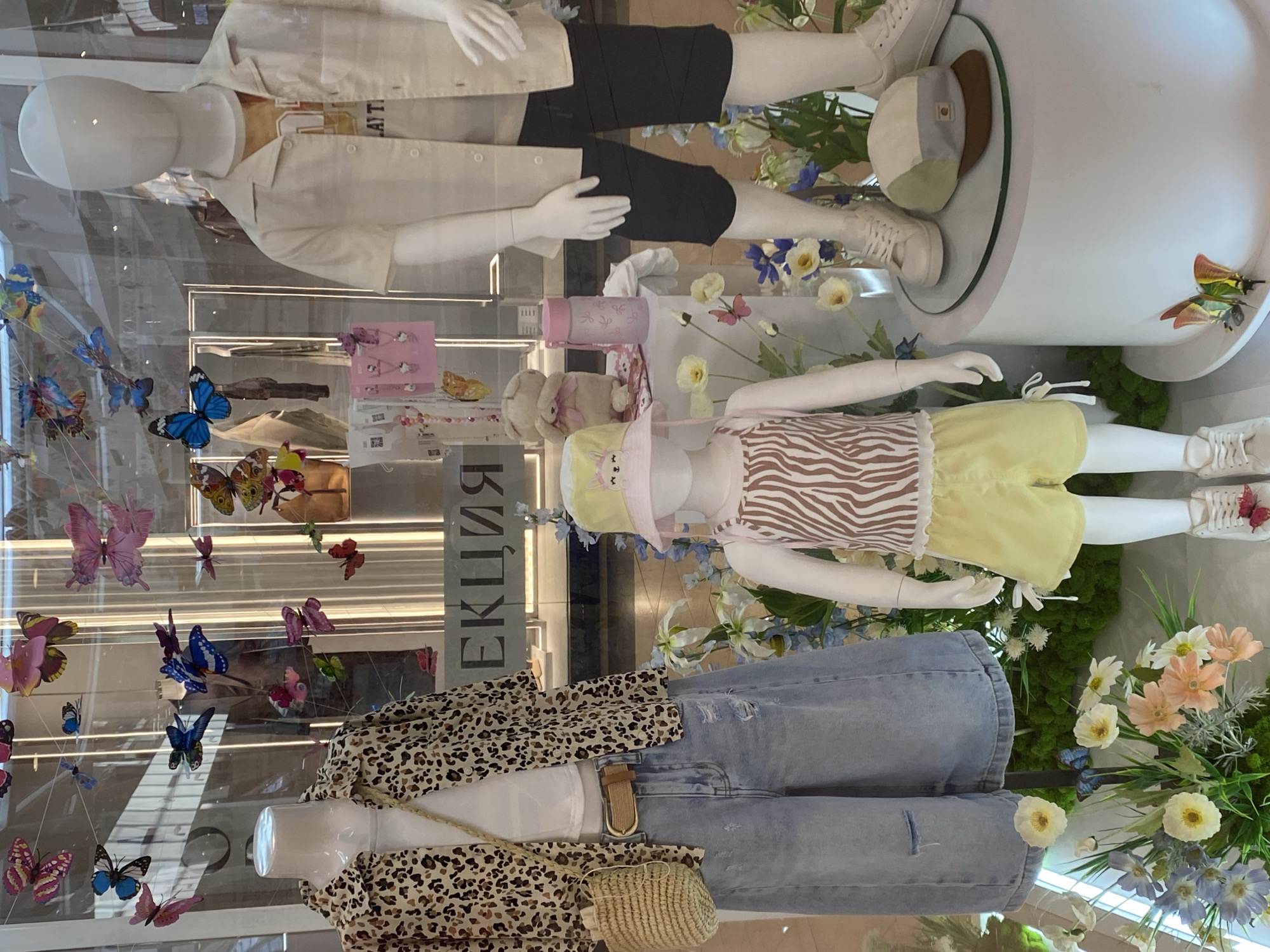 This image depicts a store window display featuring mannequins dressed in children's clothing, surrounded by artificial flowers and decorative butterflies. The display is located in Novokuznetsk, Russia.
On the left, a mannequin wears a leopard-print open-front shirt over a white t-shirt, paired with distressed light-wash denim wide-leg shorts. A woven straw bag with a brown strap hangs from its shoulder, and a brown belt cinches the waist.
In the center, a smaller mannequin is dressed in a pink and white striped tank top with a ruffled hem and yellow ruffled shorts. It wears a yellow bucket hat with a bunny face design.
To the right, a third mannequin is dressed in a light beige short-sleeved button-up shirt over a graphic t-shirt, and dark blue shorts.
The background shows a store interior with racks of clothing and signage, including the Russian word "АКЦИЯ" (AKTSIA), which translates to "SALE" or "PROMOTION." The overall scene is brightly lit, suggesting an indoor commercial setting.