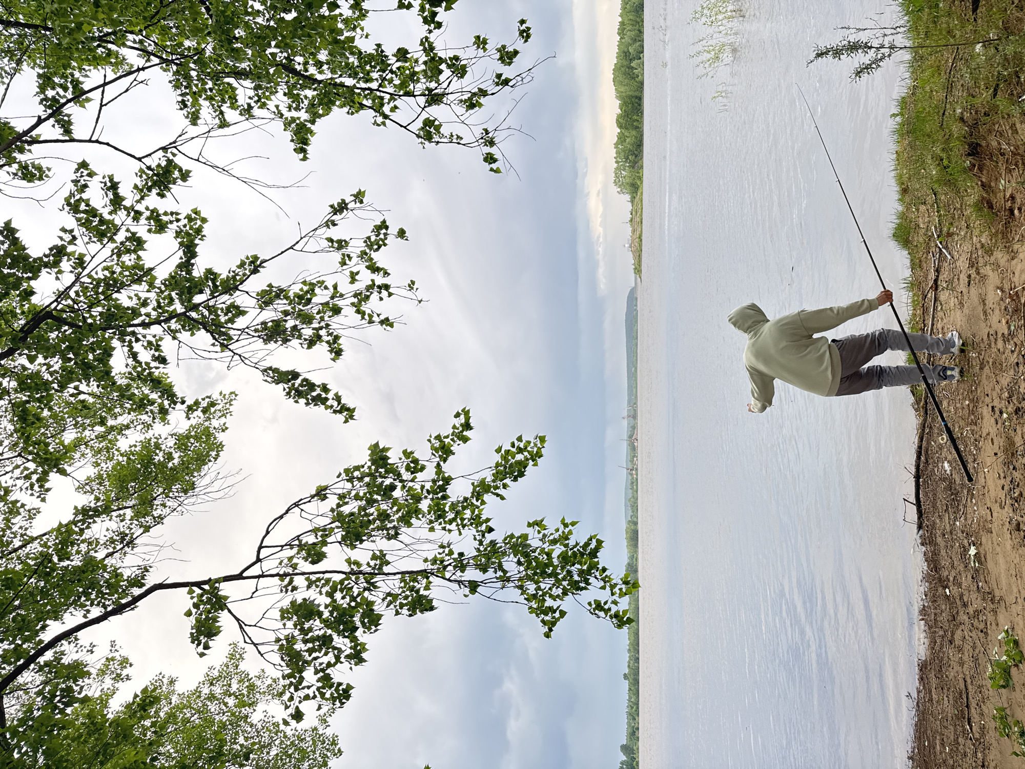 A person is fishing on the bank of a body of water. The person is wearing a light green hooded sweatshirt, gray pants, and athletic shoes. They are holding a fishing rod, casting it into the water. The water appears to be a river or lake, with a forested shoreline in the distance. There are trees with green leaves in the foreground, partially obscuring the sky which is overcast. The bank is made of dirt and is somewhat eroded. The location is possibly in or around Chaikovsky, Russia, based on the request in the prompt.
