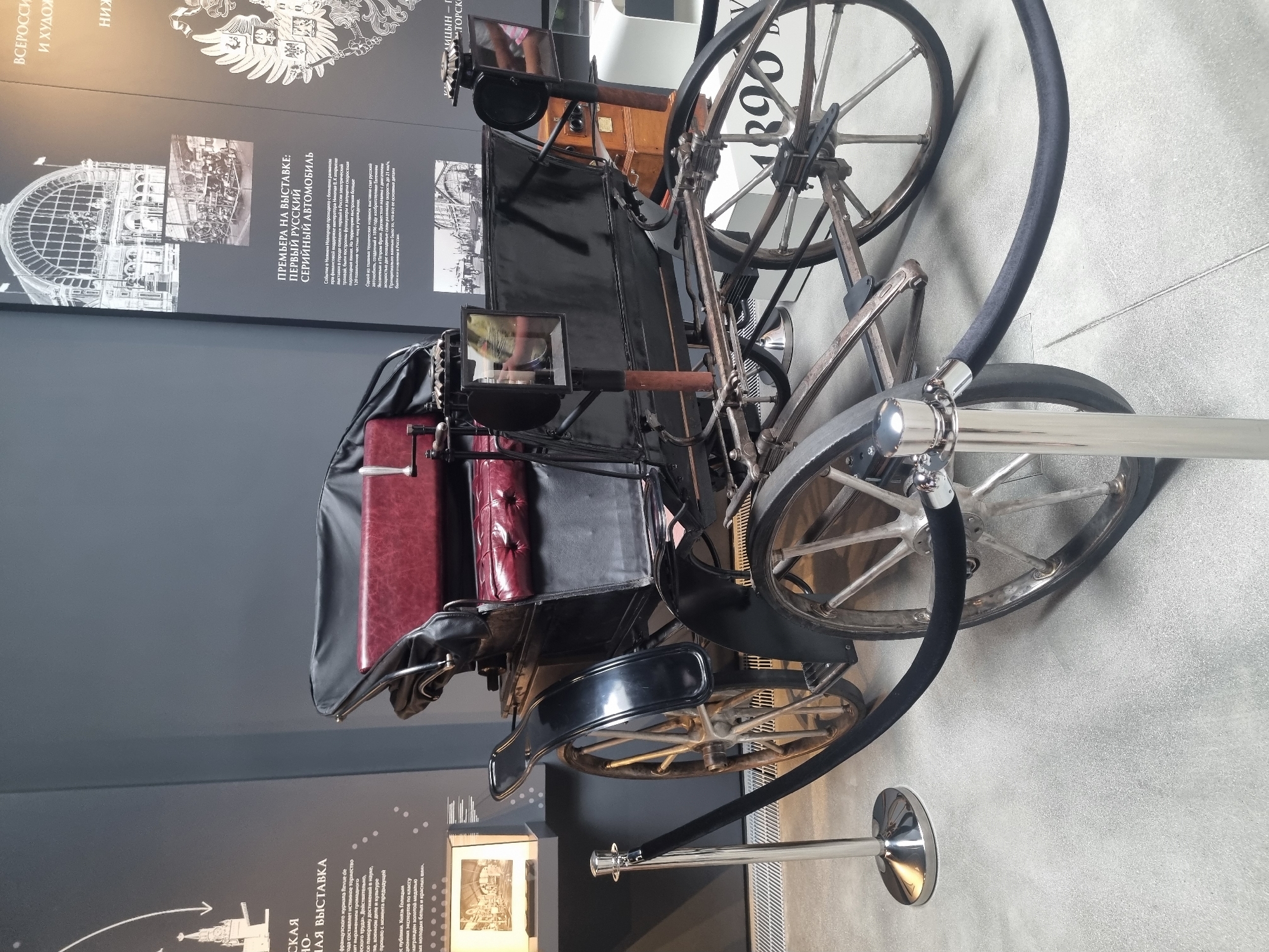 An antique black automobile with burgundy upholstered seating is displayed in a museum. The vehicle has large spoked wheels and a canvas top. It is cordoned off with stanchions and velvet ropes. The background features a dark gray wall with historical displays, including text in Russian and images of vintage buildings and people. One of the displays, in Russian, mentions "The First Russian Motor Car" and a date of "1896". The floor is light gray and tiled. The overall environment appears to be a dimly lit museum exhibit. The city of Gelendzhik, Russia is not visible in the image.